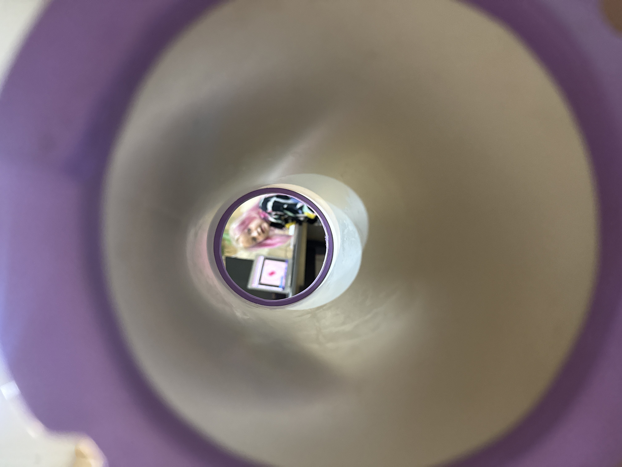 The image captures a unique perspective, looking down a wide, internal tunnel-like structure. The immediate foreground and sides of this structure are composed of smooth, curving bands of pastel purple and light, off-white or light gray, creating a sense of depth and a spiral effect.
At the far end of this tunnel, framed by a circular opening with a purple rim, a distant scene is visible. The central subject in this scene is a person with vibrant pink hair, whose face is partially visible as they appear to look towards the tunnel's opening. Due to the perspective, their features are slightly distorted.
Behind the person, on what seems to be a desk or table, several objects are discernible. A prominent feature is a tablet or screen, glowing brightly with pink or red hues, suggesting an active display. Other indistinct items, including a black and white striped object, are also present in the background.
The distant scene appears to be an indoor setting, likely a room, illuminated by artificial light, particularly from the electronic screen. The image conveys the act of viewing a subject in a different space through a distinct, enclosed passageway. There is no visible text, and the time of day or precise location cannot be determined.