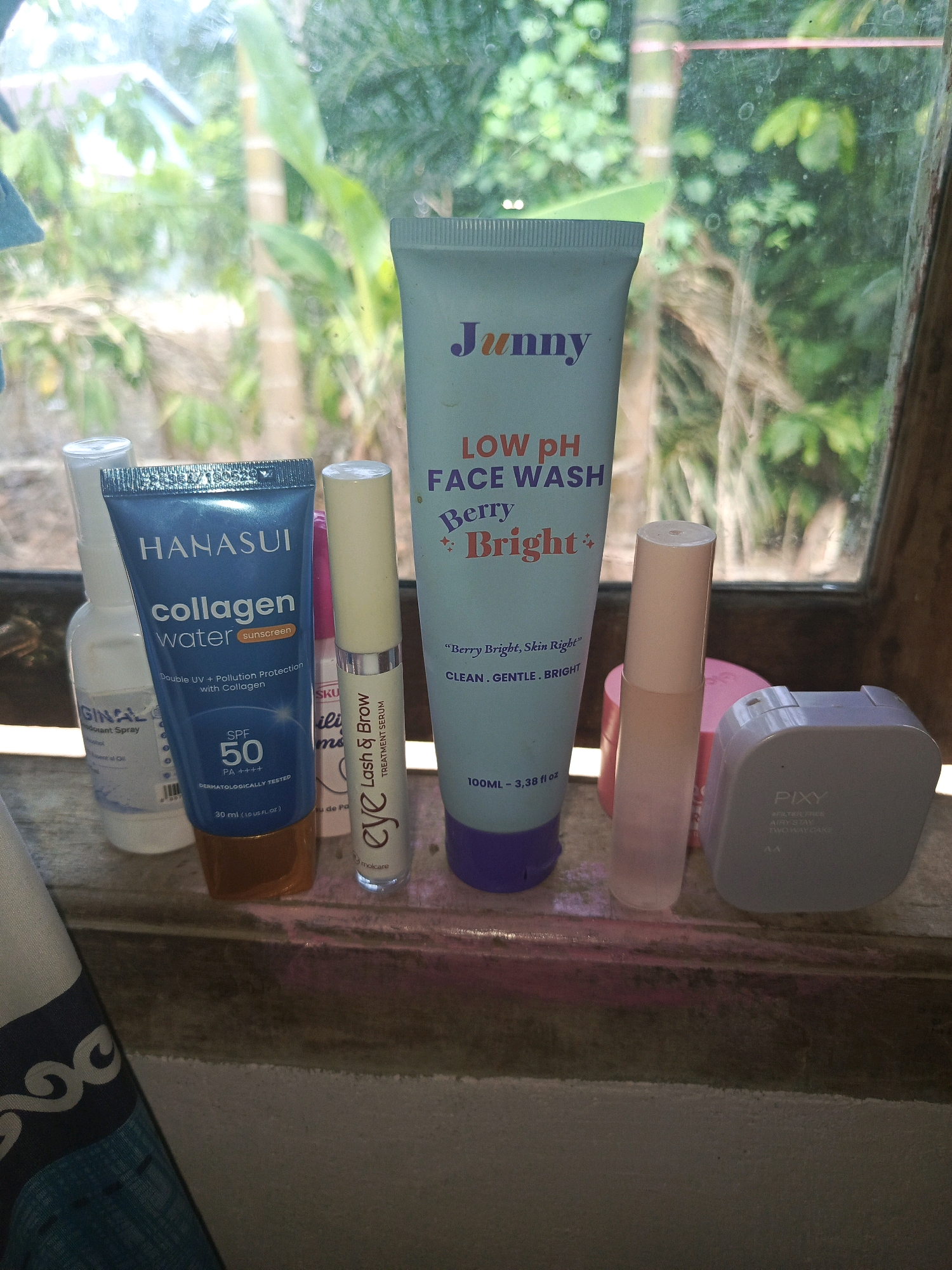 The image shows a collection of skincare products arranged on a wooden surface, likely a windowsill, with a lush green, leafy outdoor environment visible through a dirty window in the background. The lighting suggests it is daytime.
Specifically, the products visible from left to right include: a clear spray bottle with a white cap, a blue tube labeled "HANASUI Collagen Water Sunscreen SPF 50", a slender tube with a light-colored applicator labeled "eye Lash & Brow Treatment Serum", a pale blue tube labeled "Junny LOW pH FACE WASH Berry Bright", a translucent pink applicator bottle, a pink compact case, and a light gray square compact labeled "PIXY FILTER-FREE AIRY STAY TWO WAY CAKE".
The location is specified as Megang Sakti I, Indonesia, which is a rural or semi-rural area given the natural, uncultivated greenery seen outside the window. The presence of a windowsill and the type of natural backdrop suggest a residential setting or a place with natural surroundings. The overall scene depicts personal grooming items within a domestic or personal space.