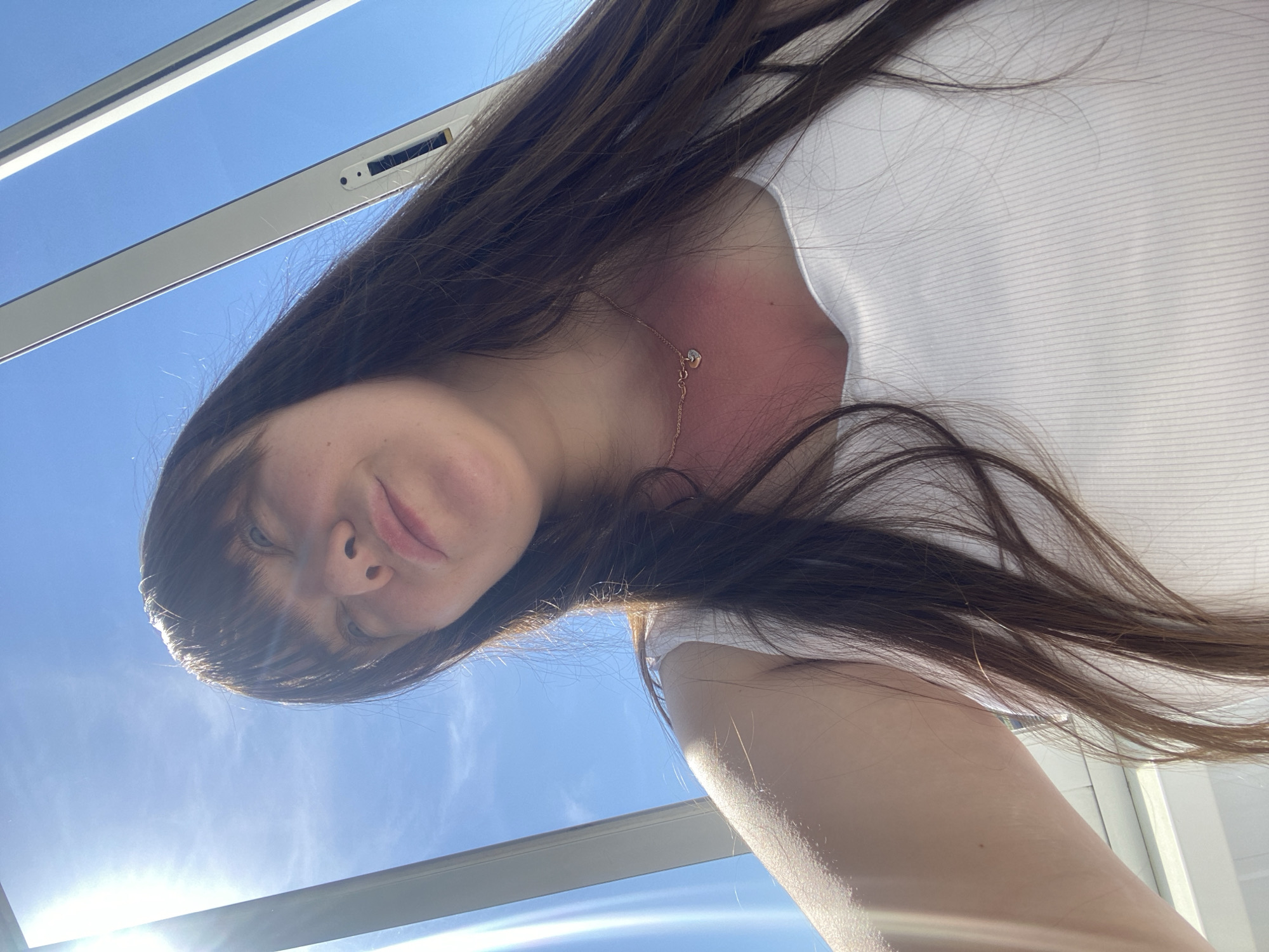 A low-angle, upward-facing photograph shows a woman with long, brown hair and fair skin. She is wearing a white top and a delicate gold necklace with a heart-shaped pendant. The background is a bright, blue sky, visible through what appears to be a skylight or window frame. The presence of a laptop is slightly perceivable due to its silver metallic finish, at the top of the frame. The city and location of Votkinsk, Russia, cannot be determined from the visual information provided.