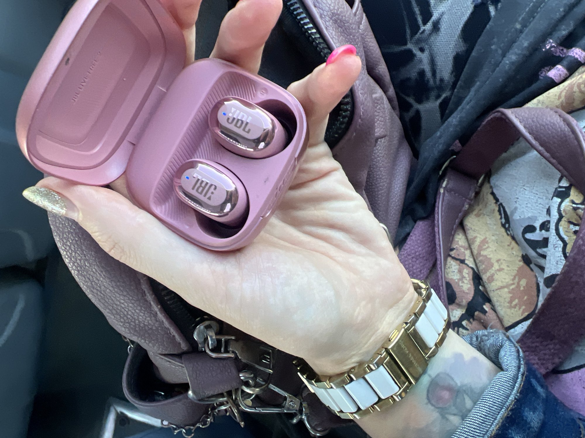 In Шолохово, Russia, a woman's hand with a glittery nail is holding a pink charging case containing two JBL wireless earbuds. The earbuds have the JBL logo on them. She is also wearing a gold and white watch. The hand is positioned over a partially visible purple leather purse with silver hardware, and a colorful fabric bag. The background is dark and out of focus.