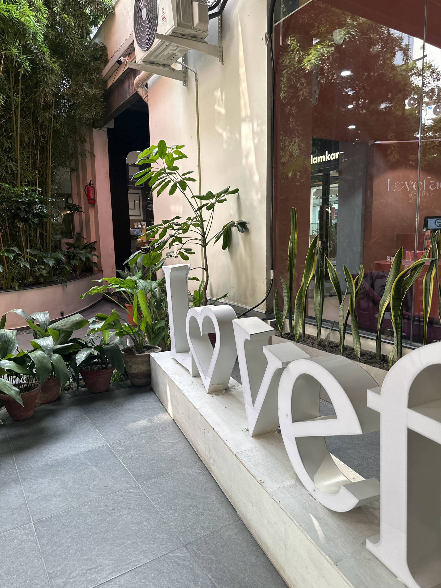 The image depicts an outdoor scene, likely in a commercial or public area, possibly in Kathmandu Metropolitan City, Nepal. In the foreground, large, white, three-dimensional letters spell out "LOVE" and then part of another word starting with "R" and ending with "R". The letters are positioned on a raised concrete platform. To the left of the letters, a variety of potted plants are arranged, including large-leafed tropical plants and a tall bamboo-like plant. A fire extinguisher is visible on a wall behind the plants. To the right of the "LOVE" sign, there is a reflective glass facade of a building, with some interior elements and signage partially visible, including the text "lamkar" and "Loveface BEAUTY". The ground is tiled with grey square tiles. An air conditioning unit is mounted on the wall above. Sunlight casts shadows across the scene, indicating it is daytime.
