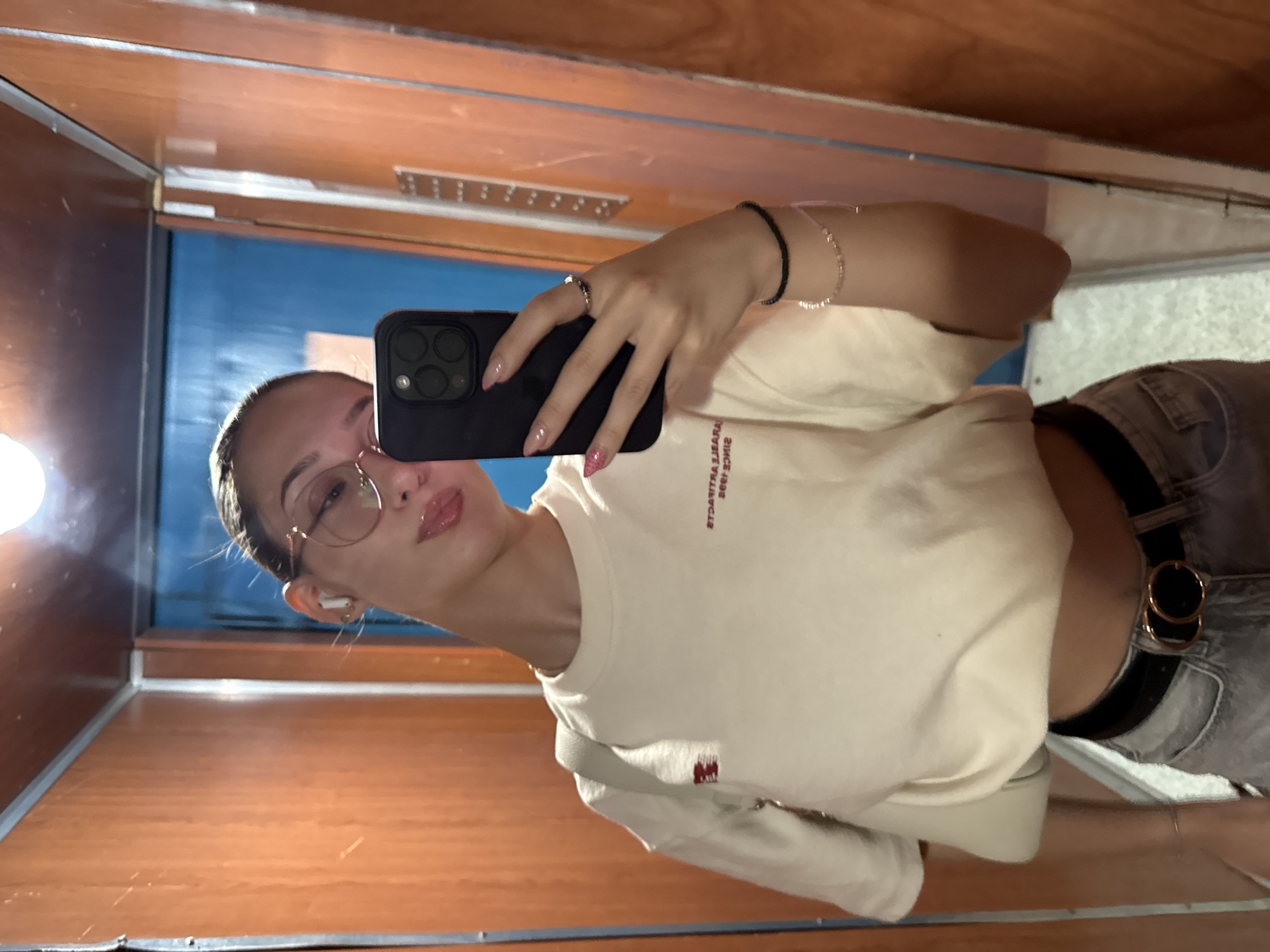 A young woman is taking a selfie in a mirror. She is wearing a cream-colored long-sleeved shirt, gray jeans, and a black belt with a double gold buckle. She has on glasses and earbuds. Her left hand is holding her phone, and her right hand is resting on her hip. She has on pink nail polish and bracelets. She appears to be in an elevator, indicated by the wooden paneling and the reflective surface of the elevator doors. The lighting is dim.