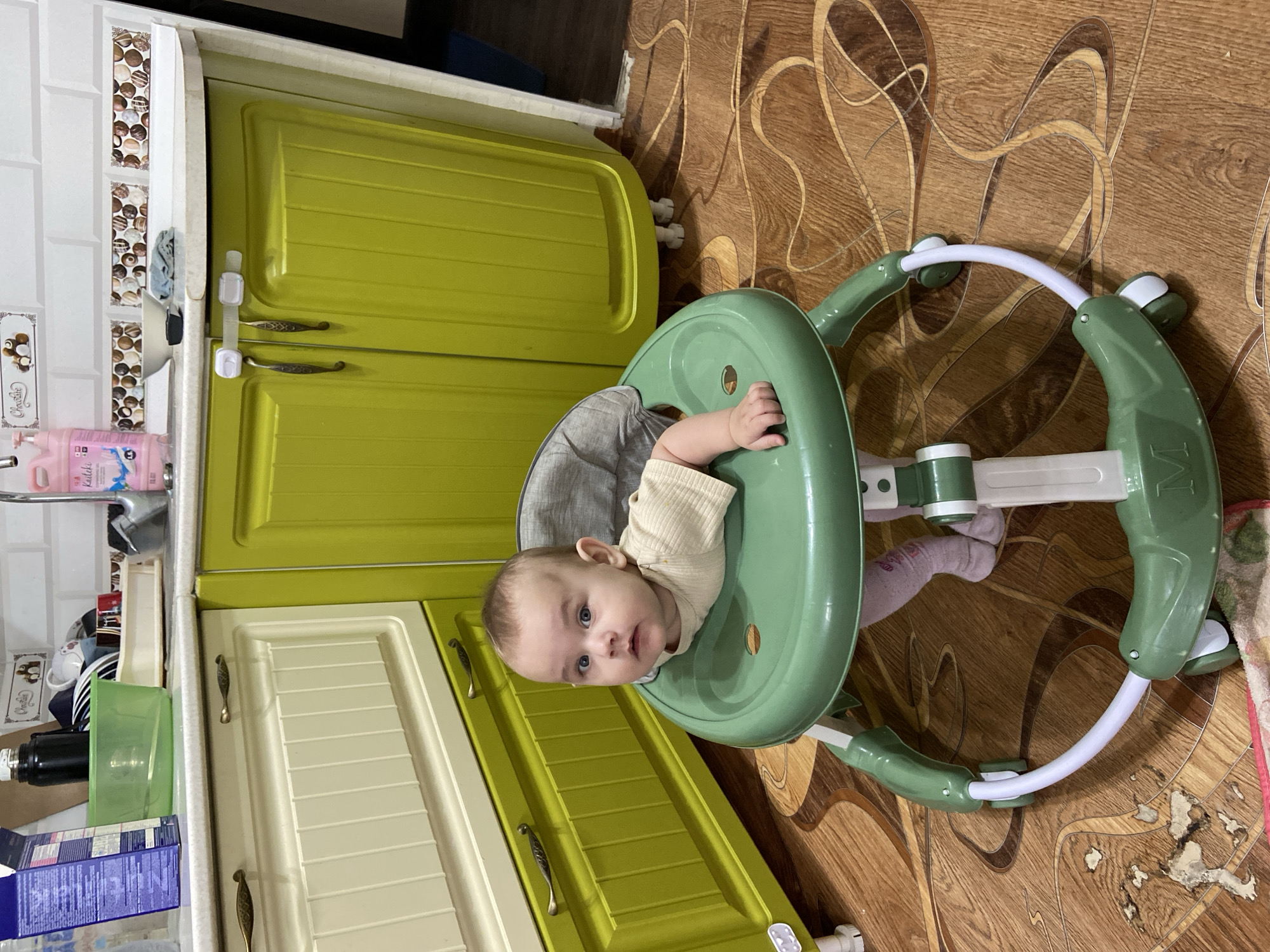 A baby sits in a green walker in a kitchen. The baby is wearing a light-colored shirt and pink socks. The walker has a gray fabric seat. The kitchen has green cabinets and a white countertop. There are various items on the countertop, including a green bowl, a black thermos, and a bottle of pink liquid. The floor is made of wood-look laminate.