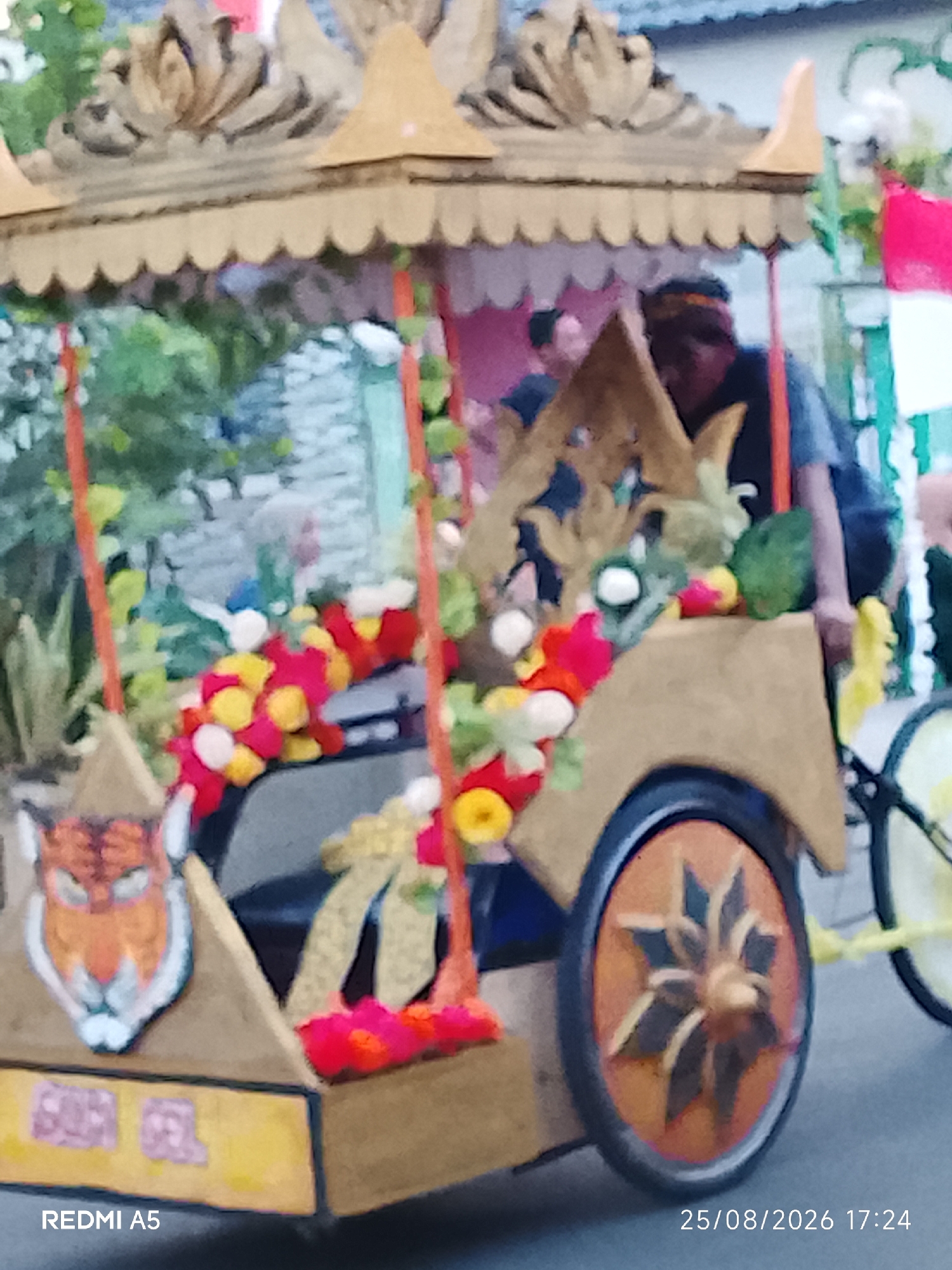 An ornate, wooden, open-air carriage, decorated with red and yellow fabric flowers and green foliage, is featured in the foreground. A stylized orange tiger face is painted on the left side of the carriage. The carriage has a patterned wooden wheel on the right. Two individuals are partially visible in the background; one wears a dark shirt and a headband, and the other wears a dark shirt. A red and white flag is positioned behind the carriage on the right. The scene appears to be set on a street, possibly during a parade or festival in Semanding, Indonesia. The bottom left of the image displays "REDMI A5" and the bottom right shows "25/08/2026 17:24".