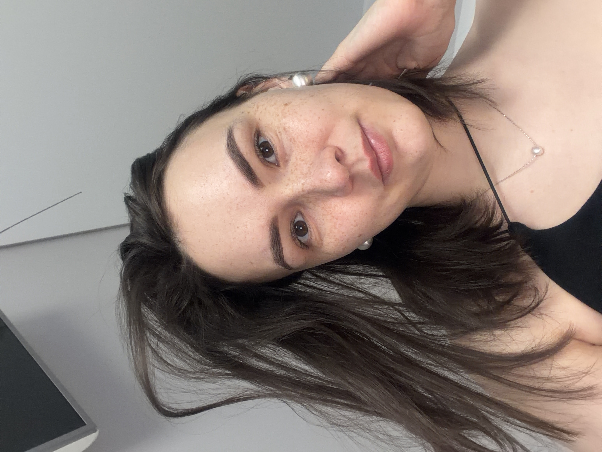 A portrait-style photograph depicts a woman with fair skin, freckles, and dark hair. Her hair is partially obscuring her neck and shoulder, which are visible in a sleeveless black garment. She is wearing pearl earrings and a delicate necklace with a single pearl pendant. Her expression is calm with a slight smile.
The background is a plain, light grey or white wall. Part of a computer monitor is visible in the lower left corner.
The location is not discernible from the provided image. It cannot be definitively determined to be in Chaikovsky, Russia.