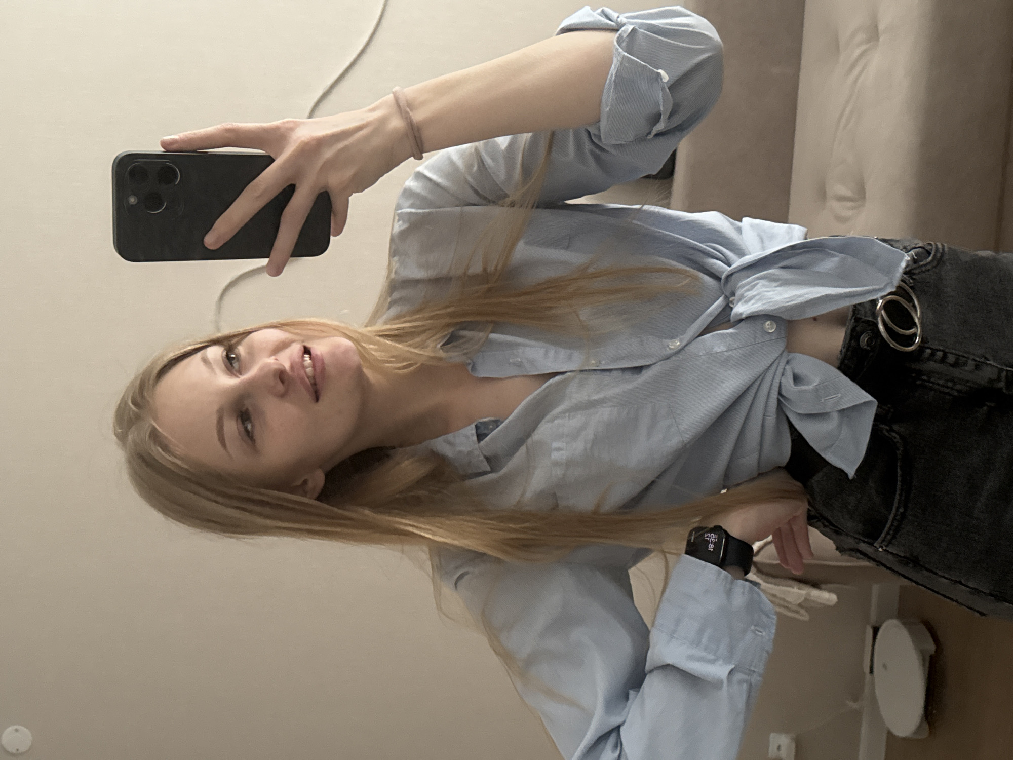 The image is a selfie taken in an indoor setting. A young woman with blonde hair is visible, wearing a light blue button-down shirt tied at the waist and dark wash jeans. She is holding a smartphone to take the picture. The background appears to be a room with a neutral-colored wall and a couch. There is a white circular object on the floor in the lower left corner, possibly a robotic vacuum cleaner. A small light fixture is visible on the wall in the upper left. The lighting suggests an indoor environment during daylight hours. There is no discernible information in the image to determine a specific location in the city of Perm, Russia.