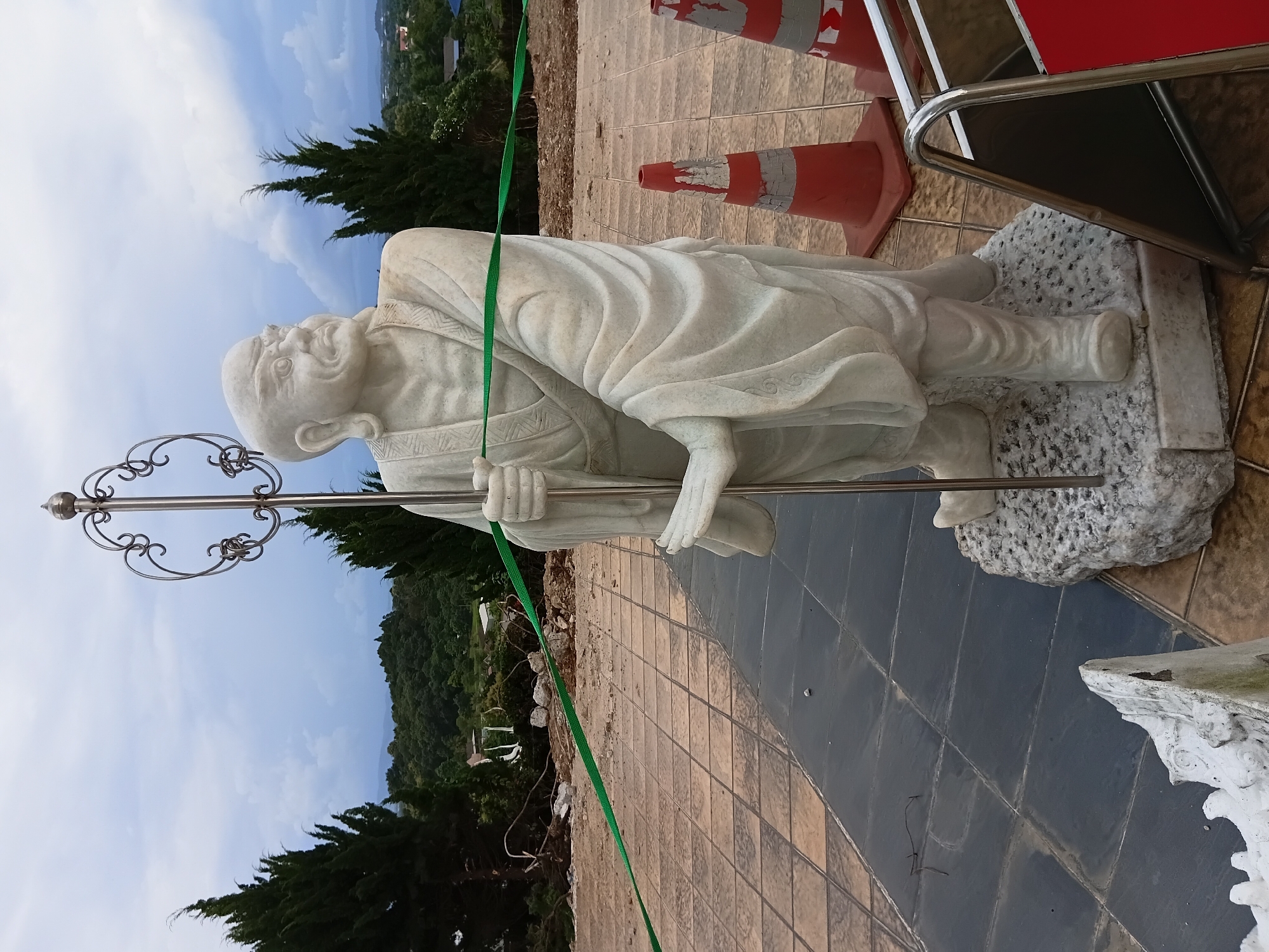 The image depicts a marble statue of a bald, elderly man wearing a robe and holding a staff with an ornate, circular metal top. The statue stands on a rough-hewn stone base. The statue is positioned outdoors on a tiled plaza, with a darker tiled path in the foreground. Behind the statue, there is a construction or landscaping site with disturbed earth, rubble, and orange traffic cones. A green ribbon is stretched across the scene. In the background, there are cypress trees, a treeline of a wooded hill, and a cloudy sky. The location is identified as Ban Lao Phatthana, Thailand, suggesting a rural or semi-rural setting within the country. The overall scene suggests an outdoor public space, possibly a temple or a monument, undergoing some form of development or maintenance.