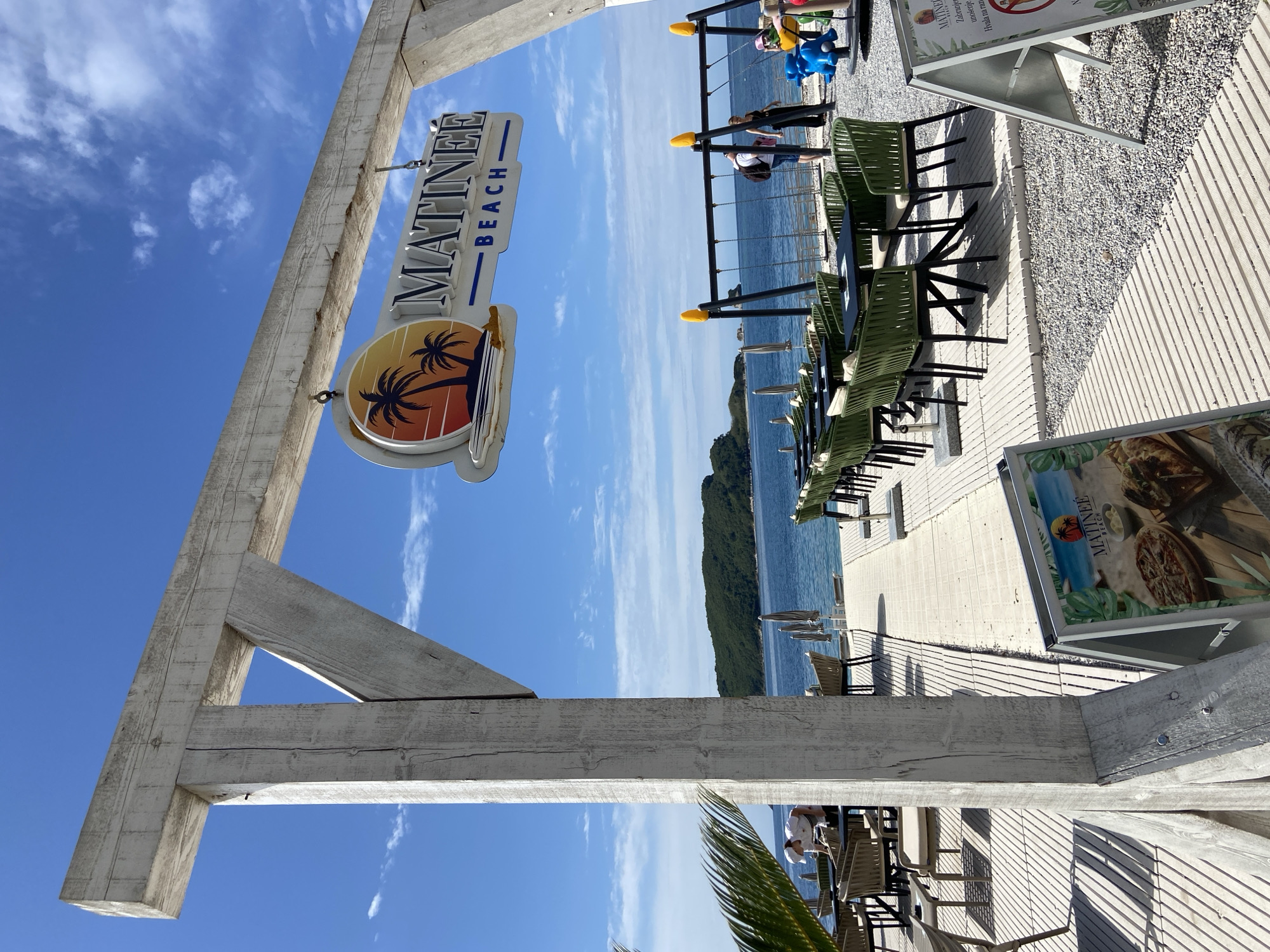 The image shows an outdoor scene at "Matinee Beach" in Budva, Montenegro. The scene features the beach sign hanging from a weathered wooden frame. Rows of green chairs and tables are positioned on a light-colored tiled walkway beside a pebble beach. A few people are visible in the background. The backdrop is a blue sea and a lush green mountainous coastline. The sky is clear with scattered clouds. In the foreground, a menu stand displays images of food.
