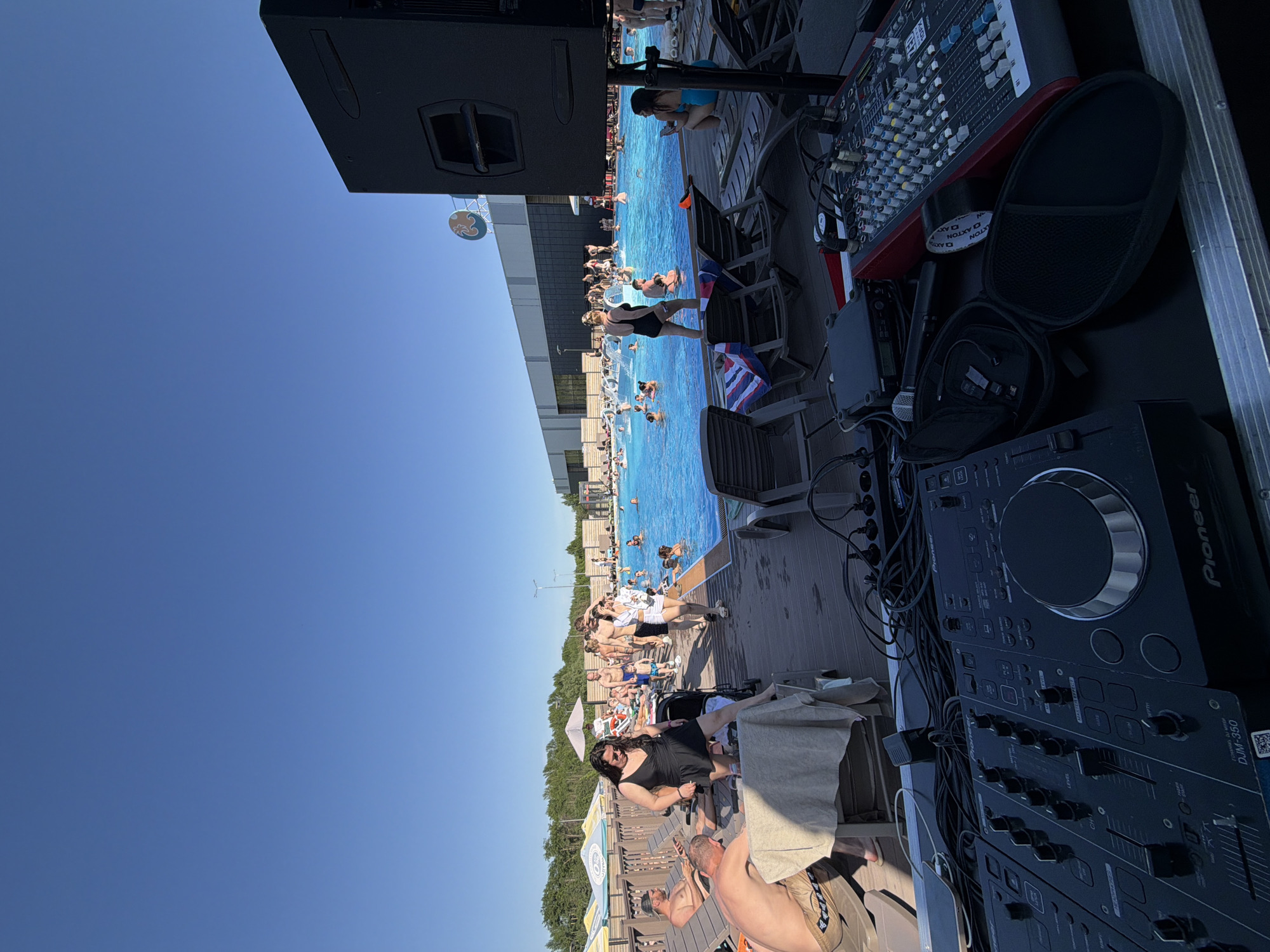 The image depicts a scene at a pool party in Kazan, Russia. A large outdoor pool is filled with people swimming and enjoying themselves. Lounge chairs line the pool deck where people are sunbathing and relaxing. A DJ setup, including a Pioneer DJM-350 mixer, is positioned at the edge of the pool area. A soundboard and other audio equipment are visible next to the mixer. The sky is clear and blue.