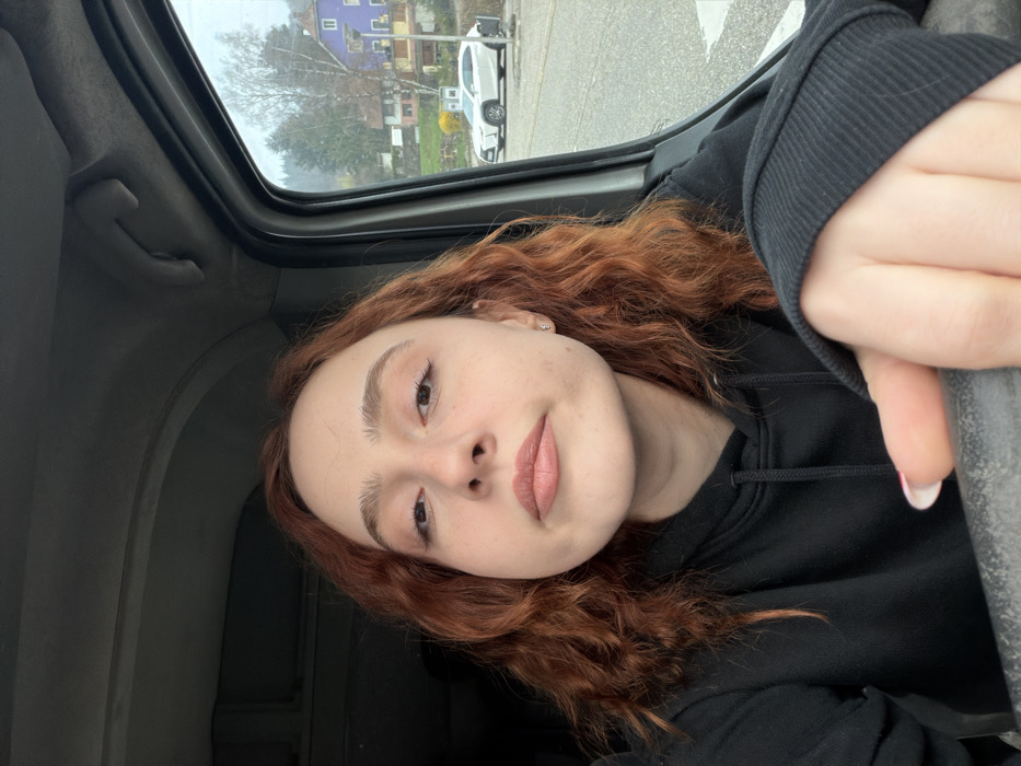 A young adult female with auburn, wavy hair and a neutral expression is captured in a selfie within the interior of a vehicle, located in Plaine, France. She has a light complexion, light eye makeup, and light-colored lipstick. A small silver stud earring is visible in her right ear. She is wearing a black hooded sweatshirt. Her right hand, featuring nails with white tips, is visible in the lower right, resting on a dark, flat surface, possibly a dashboard or door frame.
The vehicle's interior shows a light gray headliner and a hand grip above the passenger-side door frame. Outside the vehicle's window, a view of Plaine, France, reveals a street or parking area with a white Lexus SUV parked on a paved surface. The background consists of rolling hills covered with a mix of deciduous trees, some bare, and evergreens. Architectural elements include a multi-story building with a distinctive purple upper section, alongside other structures in various shades of brown and beige. Patches of yellow flowering bushes are visible near the buildings. The sky is overcast.