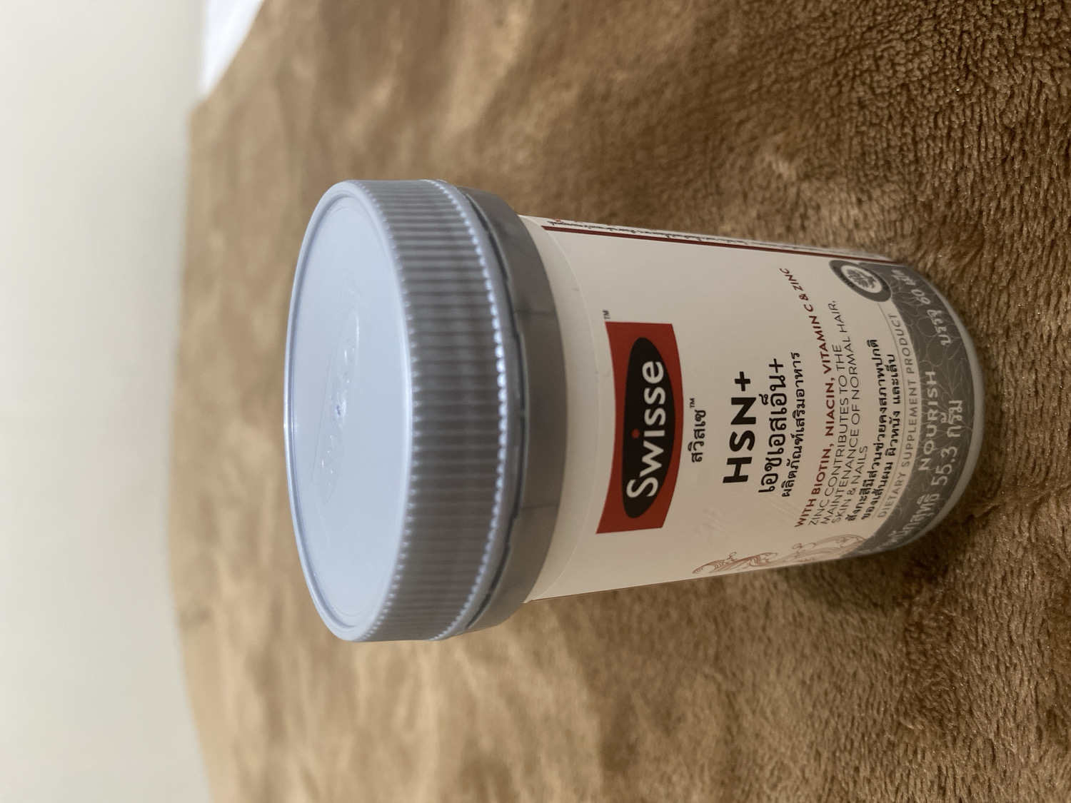 An indoor, close-up, eye-level shot features a white Swisse HSN+ dietary supplement bottle lying sideways on a textured brown surface, possibly a blanket or rug. A light-colored wall is visible in the upper left background. The bottle has a grey screw cap with ridged edges.
The label on the bottle is predominantly white with black and red text. Prominently displayed is the brand "Swisse" in red, followed by "HSN+" in large black letters. Below this, in Thai, it reads "เอชเอสเอ็น+" and "ผลิตภัณฑ์เสริมอาหาร" (Dietary Supplement).
Key information visible on the label includes:
- "WITH BIOTIN, NIACIN, VITAMIN C & ZINC"
- "ZINC CONTRIBUTES TO THE MAINTENANCE OF NORMAL HAIR, SKIN & NAILS"
- Thai text confirming the benefits for hair, skin, and nails: "สังกะสีมีส่วนช่วยคงสภาพปกติ ของเส้นผม ผิวหนัง และเล็บ"
- "DIETARY SUPPLEMENT PRODUCT"
- The word "NOURISH"
- Quantity: "บรรจุ 60" (contains 60)
- Net weight: "หนักสุทธิ 55.3 กรัม" (Net weight 55.3 grams).
The lighting suggests daytime. The image focuses on the product, providing a clear view of its branding and nutritional claims.