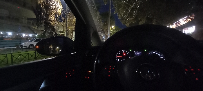 The image provides a night-time view from inside a car, looking out the front passenger side window and towards the illuminated dashboard. Outside the car, streetlights illuminate a street lined with palm trees and other foliage. In the middle ground, a white car is parked near a building with brightly lit interiors, suggesting a commercial establishment. A green metal fence separates the street from what appears to be a sidewalk or median. A small Greek flag (blue and white) is faintly visible in the background, near the building with the white car.
Inside the car, a Volkswagen (VW) steering wheel is prominent in the foreground, featuring its circular logo. The dashboard instruments are illuminated with white and green lights, showcasing a speedometer with markings up to 200 and a digital display showing status indicators, possibly including gear selection (like "D" for drive). Red/orange illuminated buttons are visible on the steering wheel and to the left of the dashboard, indicating active controls. The car's side mirror is visible on the left, reflecting some of the exterior lights.
The scene is set at night, indicated by the dark sky and artificial lighting. The overall atmosphere is calm and still, with no visible people or discernible activity beyond the car being powered on. The presence of the Greek flag aligns with the provided location context of Keratsini, Greece.