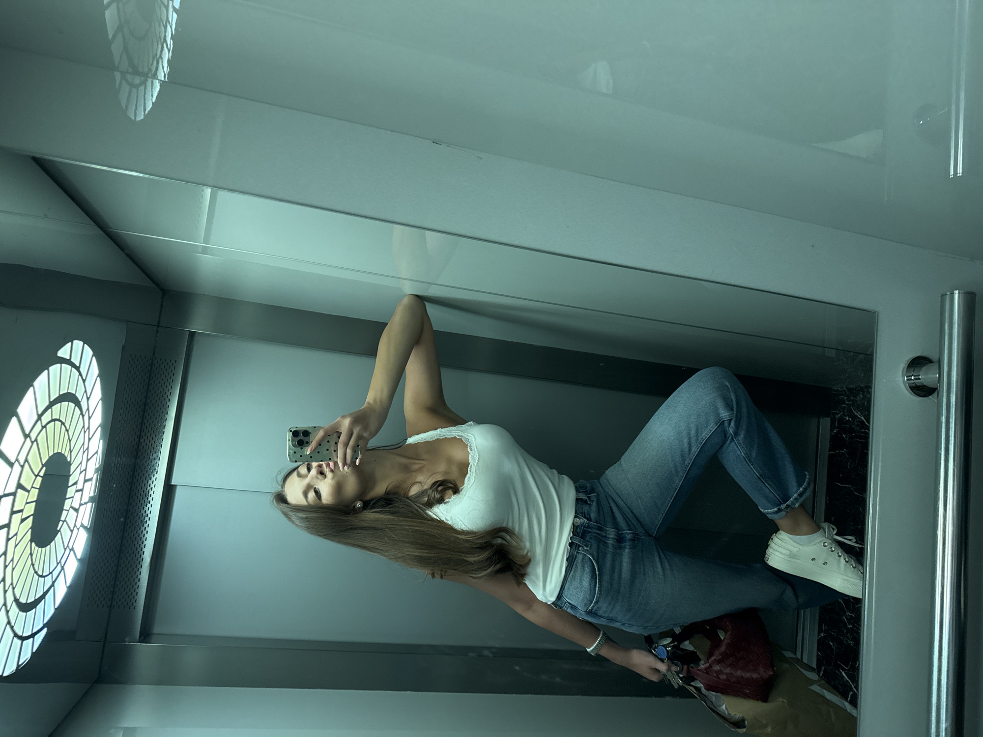 A woman is taking a selfie in an elevator. She is wearing a white tank top, blue jeans, and white sneakers. She is holding a phone in her right hand. A purse and keys are by her feet. The elevator has a large, circular light fixture on the left wall and a mirror on the right wall. Based on the light fixture design and general architectural style, the elevator appears to be located in the city of Burgas, Bulgaria.