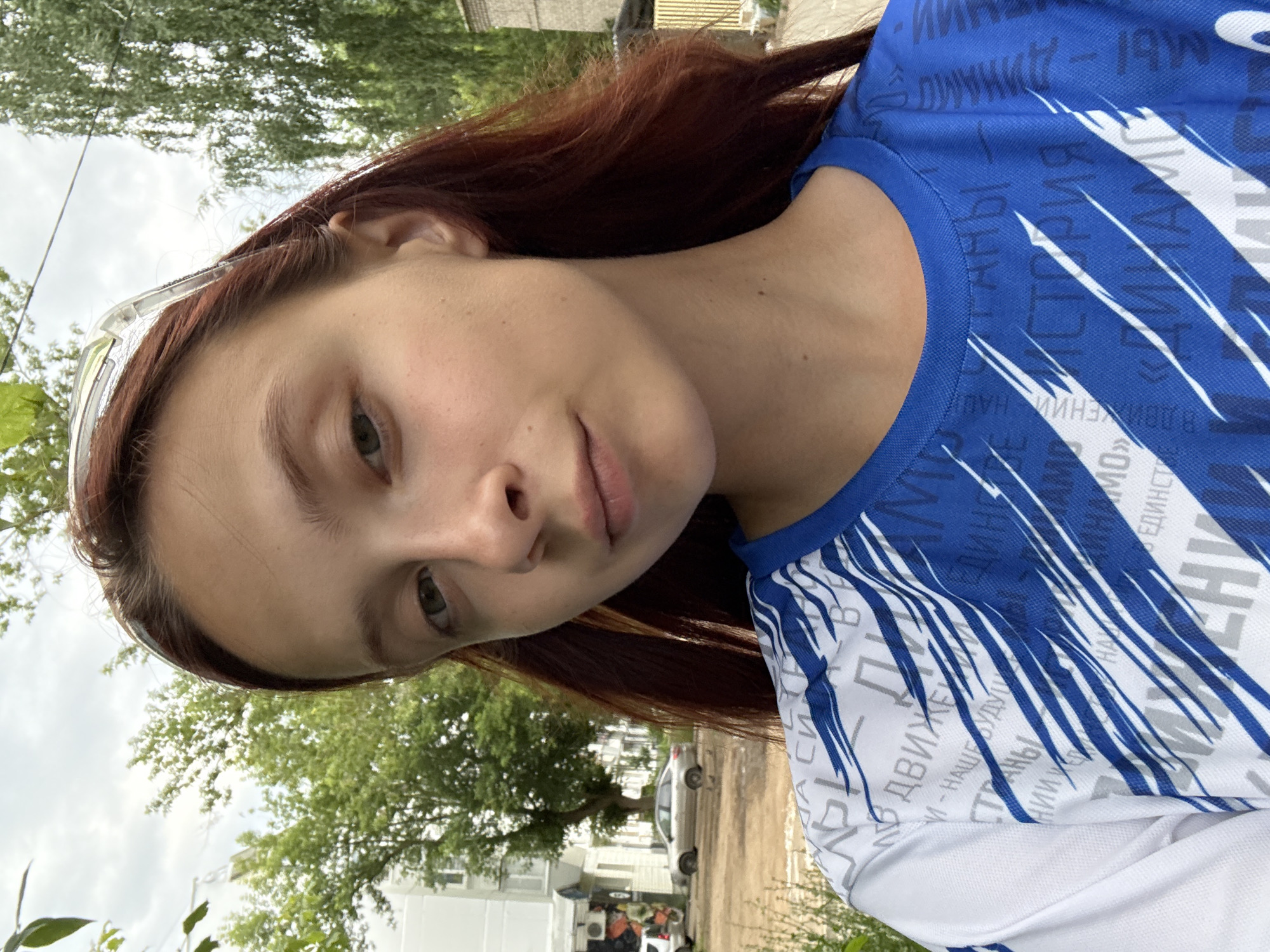 This is a vertical, close-up selfie of a young woman with reddish-brown hair and green eyes. She is wearing a blue and white patterned t-shirt with visible text in Russian. The text appears to be motivational or patriotic, with phrases like "В ДВИЖЕНИИ НАШЕ БУДУЛИ" (Our future is in motion) and "СТРАНЫ" (countries). She is outdoors, with lush green trees and a cloudy sky visible in the background. A building and a silver car are also faintly visible. The woman is looking directly at the camera with a neutral expression. The lighting suggests it is daytime, likely overcast. The setting appears to be a residential area in Chaikovsky, Russia. A clear plastic visor is perched on her forehead.