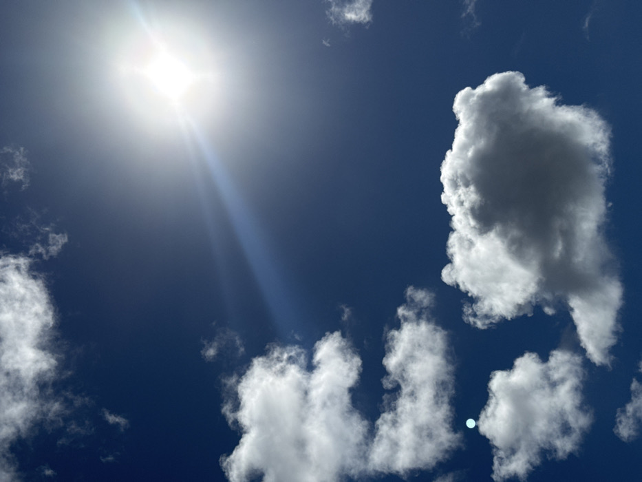 The image displays a vibrant, clear blue sky dotted with scattered white clouds, illuminated by a brilliant sun. The sun is positioned in the upper left quadrant of the frame, appearing as an intensely bright, almost star-like light source, with noticeable lens flares or sunbeams extending diagonally downwards and to the right, casting a bright sheen across that area of the sky. The sunlight is strong, suggesting a clear and bright day.
The sky is a deep, rich blue, contrasting sharply with the bright white of the clouds. There is a prominent, large, and dense cumulus cloud occupying a significant portion of the upper right side of the image. Its edges are bright white where directly hit by sunlight, while its interior shows some grayish tones, indicating its volume and depth. Below this large cloud, and extending across the bottom center and lower left, are several smaller, fluffier white clouds, also brightly lit. Some wispy cloud formations are also visible closer to the sun in the top left.
A subtle, small, bluish-white circular artifact or reflection is visible near the bottom center of the image, amidst the clouds, which could be a lens flare. The overall impression is one of a beautiful, sunny day with fair-weather clouds. No text is visible in the image.