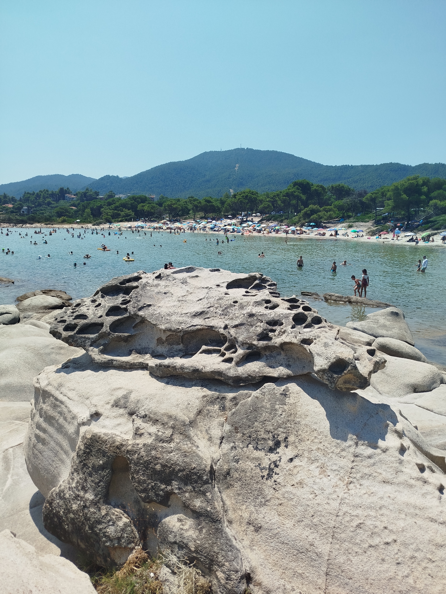 The image depicts a sunny day at a crowded beach in Sithonia Municipal Unit, Greece. In the foreground, large, weathered rocks with natural formations dominate the scene. The middle ground shows the clear, blue water filled with numerous people swimming, wading, and relaxing on inflatable objects. A sandy beach is visible in the background, lined with trees and more people sunbathing under umbrellas. Lush green hills form the backdrop of the scene. The overall atmosphere is one of a vibrant, summer beach day.