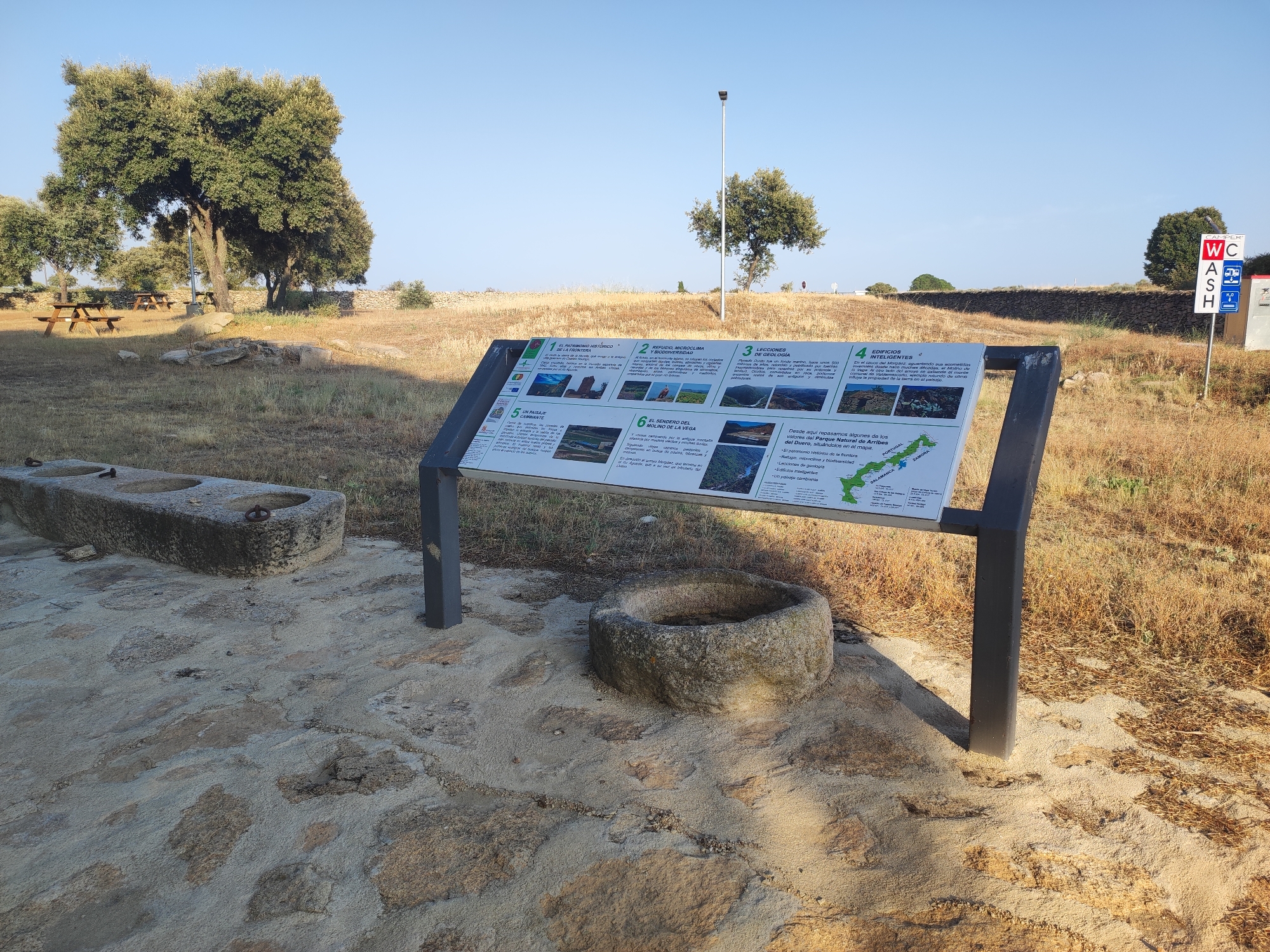 An outdoor informational display is situated in a dry, grassy landscape under a clear blue sky. The display, mounted on black metal poles, presents a large graphic with multiple sections, each containing text and images. To the left of the display is a long, rectangular stone trough with two indentations, possibly for watering livestock, and to the right is a circular stone basin. In the background, several trees are visible, including a large, mature tree to the left. A stone wall runs horizontally across the middle ground. Further in the distance, a sign with "WC" and "CASH" is visible. The ground is covered with dry grass and scattered stones, with a paved area of irregular stone slabs in the foreground. The scene suggests a historical or natural park area.
