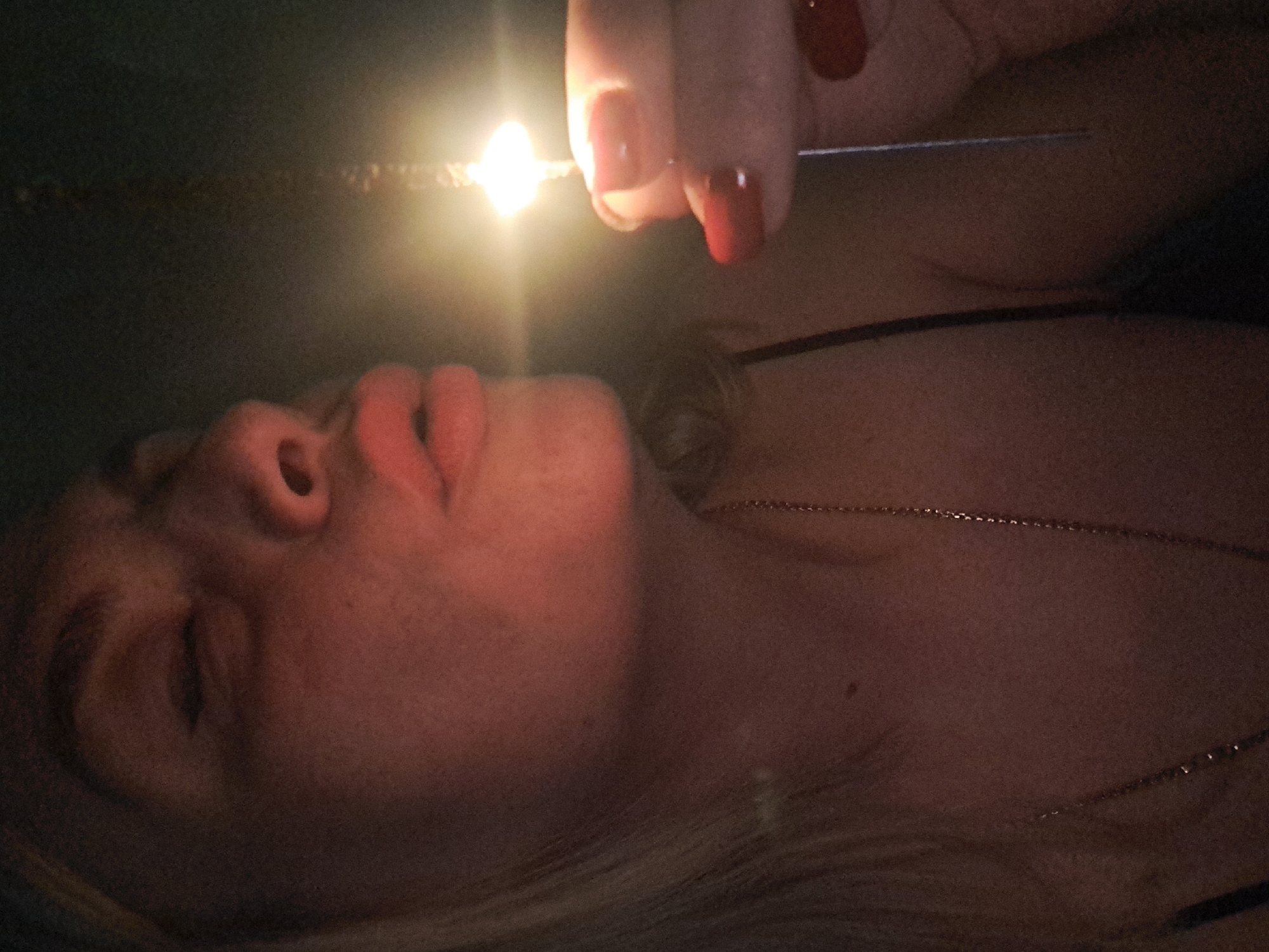 A person is lying on their back, with their face illuminated by a bright sparkler held above them. The person's face is turned upwards, with their eyes partially closed. A hand holding the sparkler is visible in the upper part of the frame. The sparkler is emitting a bright light. A thin necklace is visible on the person's chest. The background is dark and indistinct. The location in Elektrostal, Russia, is not discernible from the image.