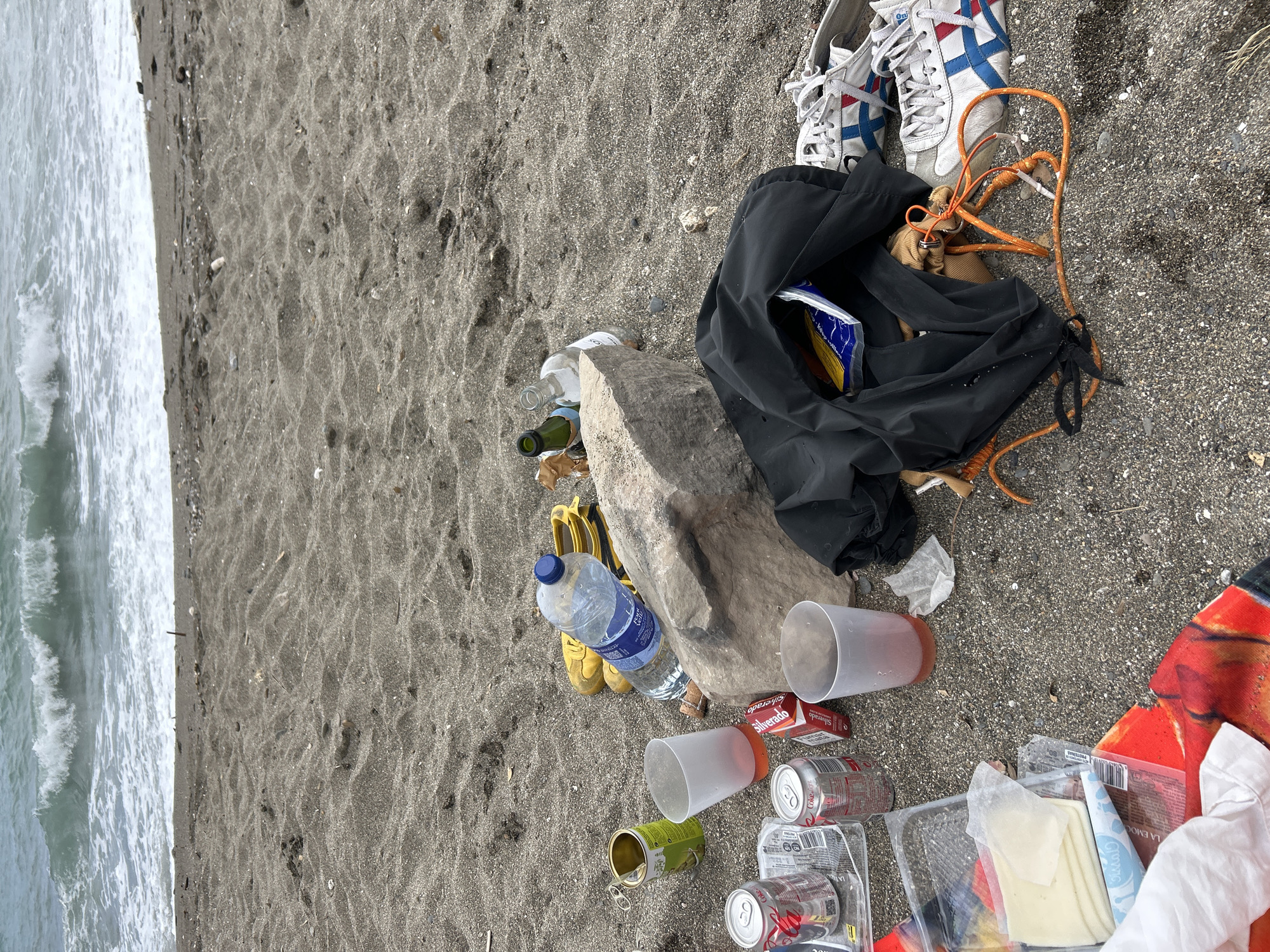 The image captures a significant amount of litter discarded on a dark sand beach next to the ocean. The scene is dominated by various human-made objects scattered across the foreground, with the ocean and breaking waves visible on the left side of the frame.
In the center and right, a collection of items includes a black fabric bag, possibly a drawstring bag, with an orange rope attached. Next to it are a pair of white sneakers with blue and red stripes, and further back, yellow flip-flops are partially visible. A large, light-colored rock sits among the debris.
Surrounding these items is an array of discarded food and drink containers: several empty glass bottles (clear and green), a large plastic water bottle, multiple empty plastic cups (some frosted, some with reddish liquid residue), numerous empty aluminum soda cans (some clearly marked "Coca-Cola"), an open tin can, and various plastic food packaging, including a container of cheese slices and other wrappers. A "Silverado" cigarette pack is also visible. Crumpled paper and plastic waste are interspersed throughout the litter.
The activity suggested by the scene is the aftermath of a beach gathering or casual visit, leaving behind a substantial amount of garbage. The bright, natural lighting indicates it is daytime, likely a sunny day. The location cues are clearly a beach environment with dark, coarse sand and ocean waves. No people are visible.