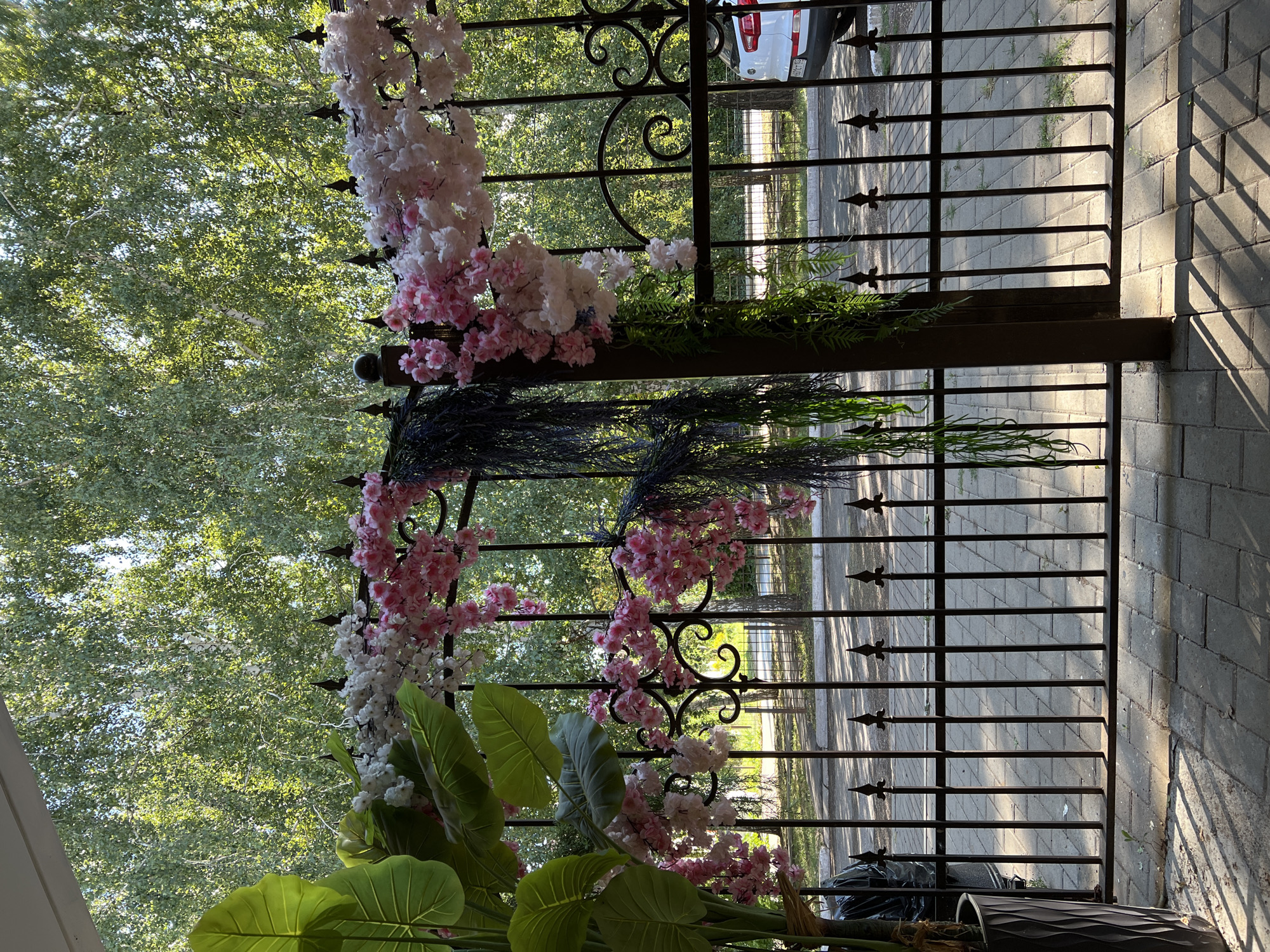 The image depicts an outdoor scene with a black wrought-iron fence in the foreground. The fence is decorated with artificial floral arrangements, featuring pink and white flowers, and cascading greenery. To the left, a large potted plant with broad green leaves is partially visible. Beyond the fence, a paved area with sunlight casting shadows is visible, and further back, a white car is parked. The background consists of a dense canopy of green trees, suggesting a park or a wooded area.
The scene is located in the city of Salavat, Russia. Specifically, the fence and floral decorations are likely part of a business or event space, possibly an outdoor cafe or restaurant, or an entrance to a private property, given the decorative elements and the presence of a parked vehicle. The lighting suggests it is daytime, possibly late afternoon due to the angle of the shadows. No people are visible in the image.