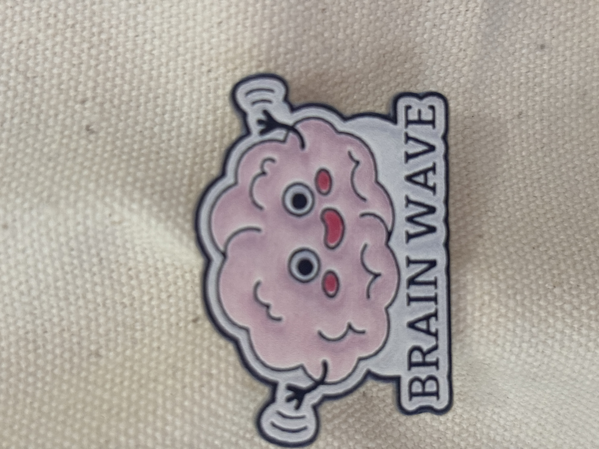 The image features a close-up of a whimsical enamel pin or sticker depicting a stylized cartoon brain. The brain is predominantly pink, with a black outline and a thin light gray border. It has a playful facial expression, featuring two large, dark, circular eyes with white highlights, a smaller red oval shape that could be a nose or another eye, and a tiny red mouth-like feature. Two short, black, stick-like arms extend from the sides of the brain, each ending in a curled, spring-like or wave-like shape, suggesting movement.
To the right of the brain character, the words "BRAIN WAVE" are spelled out vertically in dark gray capital letters against a rectangular light gray background, also outlined in black. The entire pin is set against a textured, light beige or off-white woven fabric, possibly canvas, which has a distinct, small-dot pattern visible throughout. The fabric background is slightly out of focus, emphasizing the sharp details of the pin. The design creatively illustrates the idiom "brainwave," representing a sudden idea or flash of inspiration. The photograph appears to be taken in a well-lit environment. While the location context is Sofia, Bulgaria, there are no specific visual cues within the image itself to indicate this.
