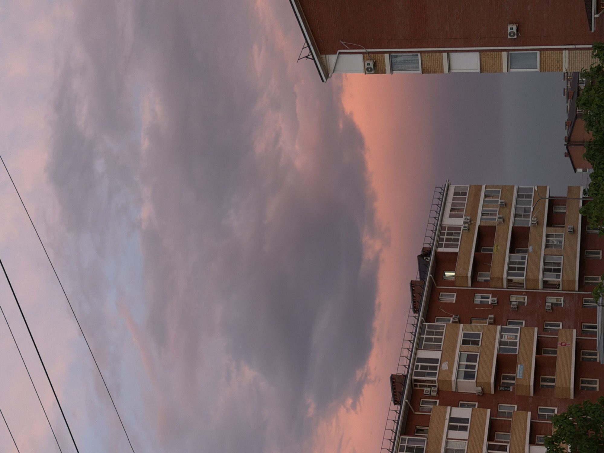 A low-angle, portrait-oriented shot reveals a building on the right side of the frame, constructed of red brick with beige accents around the many windows and air conditioning units attached to the facade. Adjacent to this building is a portion of another red brick building, visible in the upper right corner. In the background, a cloudy sky fills the majority of the frame with hues of pink, purple, and gray. Several power lines stretch diagonally from the bottom left corner toward the upper right, adding a grid-like element to the composition. The photograph captures the city of Rossiyskiy, Russia.