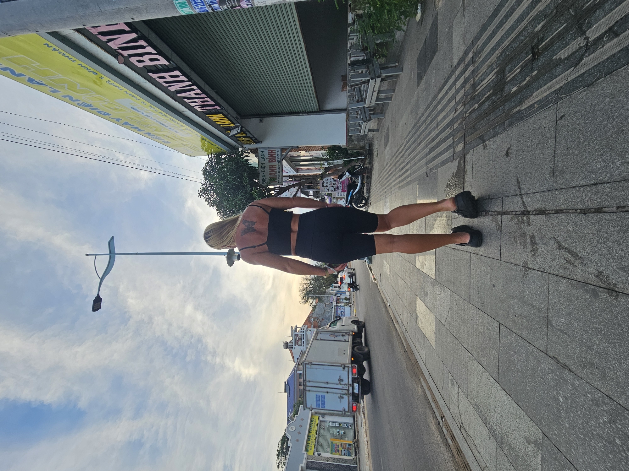 A woman with blonde hair and a butterfly tattoo on her back walks away from the camera down a sidewalk in Phu Thuy Ward, Vietnam. She is wearing a black sports bra and black bike shorts. The sidewalk is paved with gray stone tiles. To the right of the sidewalk is a building with a large yellow sign in Vietnamese and a green metal security gate. There are several wooden benches in front of the building. To the left of the sidewalk is a street with a white truck, a motorcycle, and a car driving on it. The sky is cloudy and a street lamp is visible in the distance. The overall scene suggests a street in a commercial area.