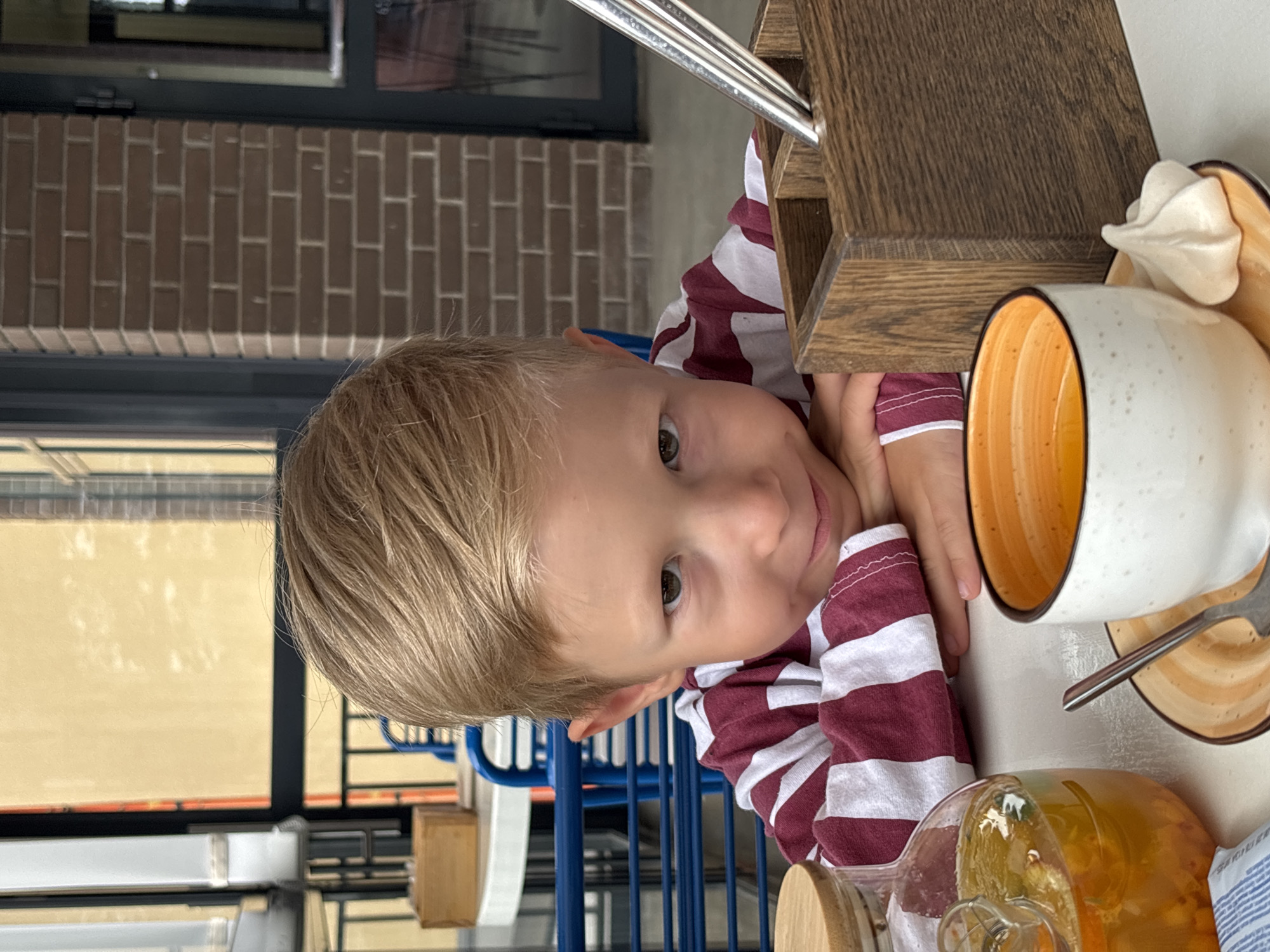 In a brightly lit outdoor cafe, likely in the city of Chaikovsky, Russia, a young boy with blond hair and a striped maroon and white shirt rests his chin on his folded hands. He gazes towards the viewer with a slight smile. In front of him, a table is set with a clear glass carafe filled with an amber beverage containing orange slices and berries, a white and orange ceramic mug with a spoon resting on its saucer, and a wooden holder containing silverware. The background features a brick wall and a glimpse of a windowed building.