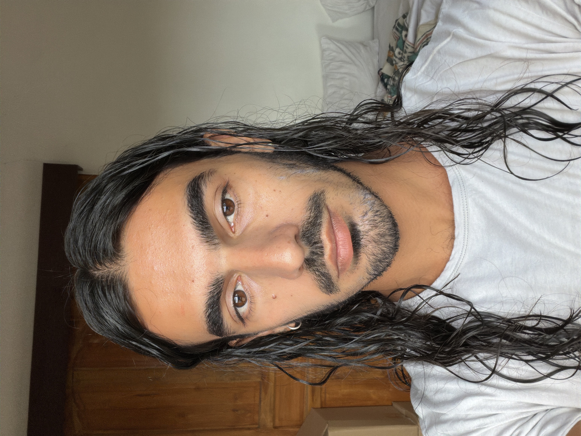 A person is facing the camera. The person has long, dark hair that appears wet, brown eyes, and a dark beard. They are wearing a white t-shirt. The background includes a light-colored wall, a white pillow, and a wooden surface. The image was taken in North Kuta, Indonesia.