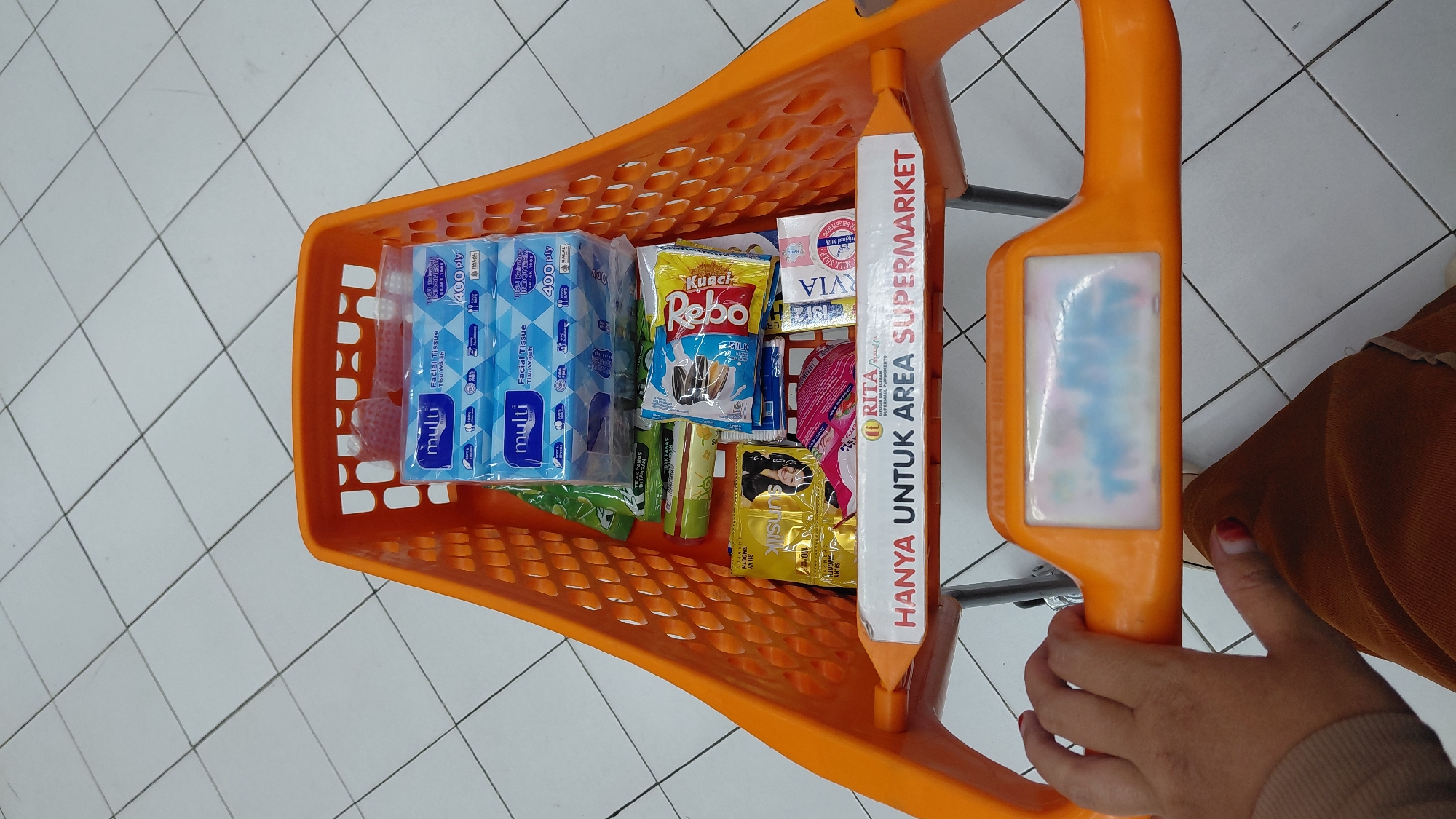 An overhead shot captures the contents of an orange plastic shopping cart in a tiled supermarket aisle in Purwokerto, Indonesia. The cart is filled with various grocery items, including several packs of "multi" brand facial tissues, a green packaged item with red text indicating "TIDAK PANAS DILARANG," a tube-like item, a yellow packet with "sunsilk" branding, and a red and yellow packet of "Rebo kuaci" sunflower seeds. Additionally, there is a blue and white package labeled "VIA" and a small white box. A prominent red and white sign at the front of the cart reads "HANYA UNTUK AREA SUPERMARKET" which translates to "ONLY FOR SUPERMARKET AREA." In the lower left corner, a person's hand, with painted fingernails, rests on the cart's handle, and their brown corduroy pants are visible. The floor is tiled in a white grid pattern, characteristic of a commercial establishment.
