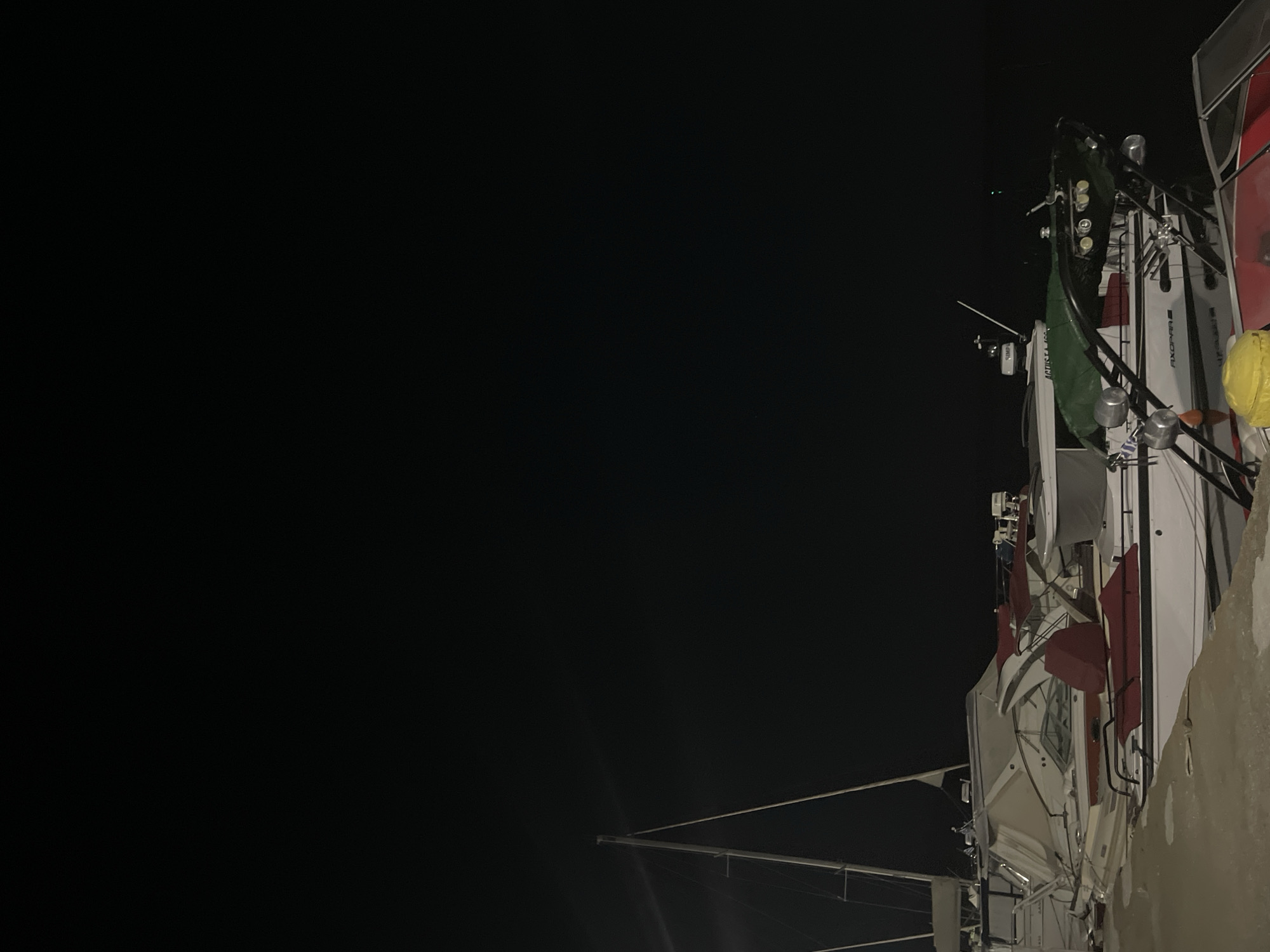 At night in Limenas Avdiron, Greece, several boats are docked at a marina. The foreground shows a concrete pier. A white and red motorboat with a green cover over part of its deck is prominently featured, with a yellow object on its deck. Behind it, another boat with a white hull and red seating is partially visible. Further back, a sailboat with tall masts stands out against the dark sky. The overall scene is illuminated by artificial lights, casting reflections on the water and highlighting the boats. The sky is dark and clear, with no visible celestial bodies. The water appears calm.