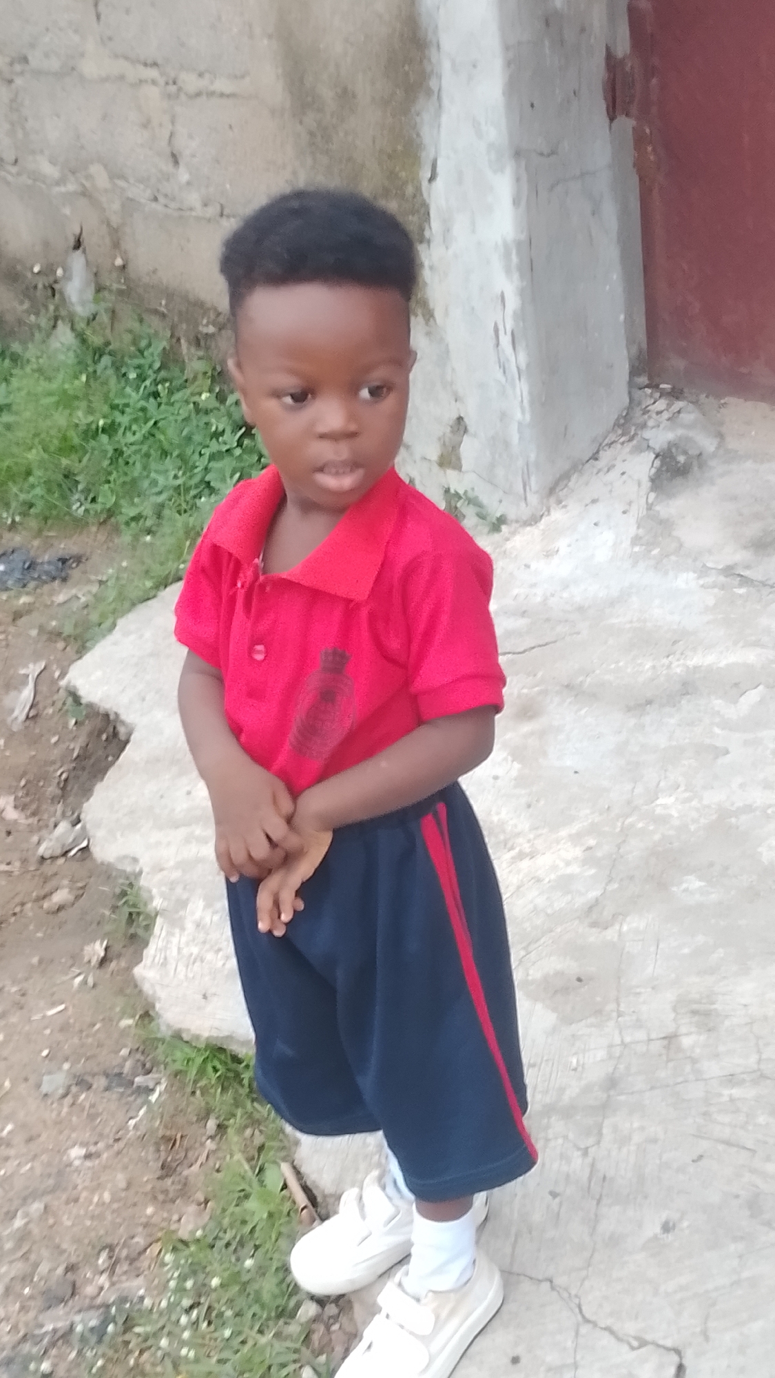 A young child stands outdoors in Lokoja, Nigeria, captured in a full-body shot. The child has dark skin, short black hair, and is wearing a red short-sleeved collared polo shirt with a dark, indistinct emblem or graphic on the left chest. They are also dressed in dark blue shorts with a vertical red stripe down each side, white socks, and white shoes with velcro closures. The child stands with hands clasped together in front, looking slightly to the right of the frame with a neutral expression and mouth slightly open.
The background features a rough, light-colored concrete wall or structure behind and to the child's right, showing signs of weathering and dark patches. To the far right, a glimpse of a reddish-brown surface, possibly a door or part of a building, can be seen. The ground beneath the child is a mix of concrete pavement and patches of dirt with sparse green vegetation, particularly prominent on the left side of the image. The lighting indicates it is daytime, likely natural light without strong direct sunlight. No discernible text is visible in the image, apart from the blurry graphic on the child's shirt.