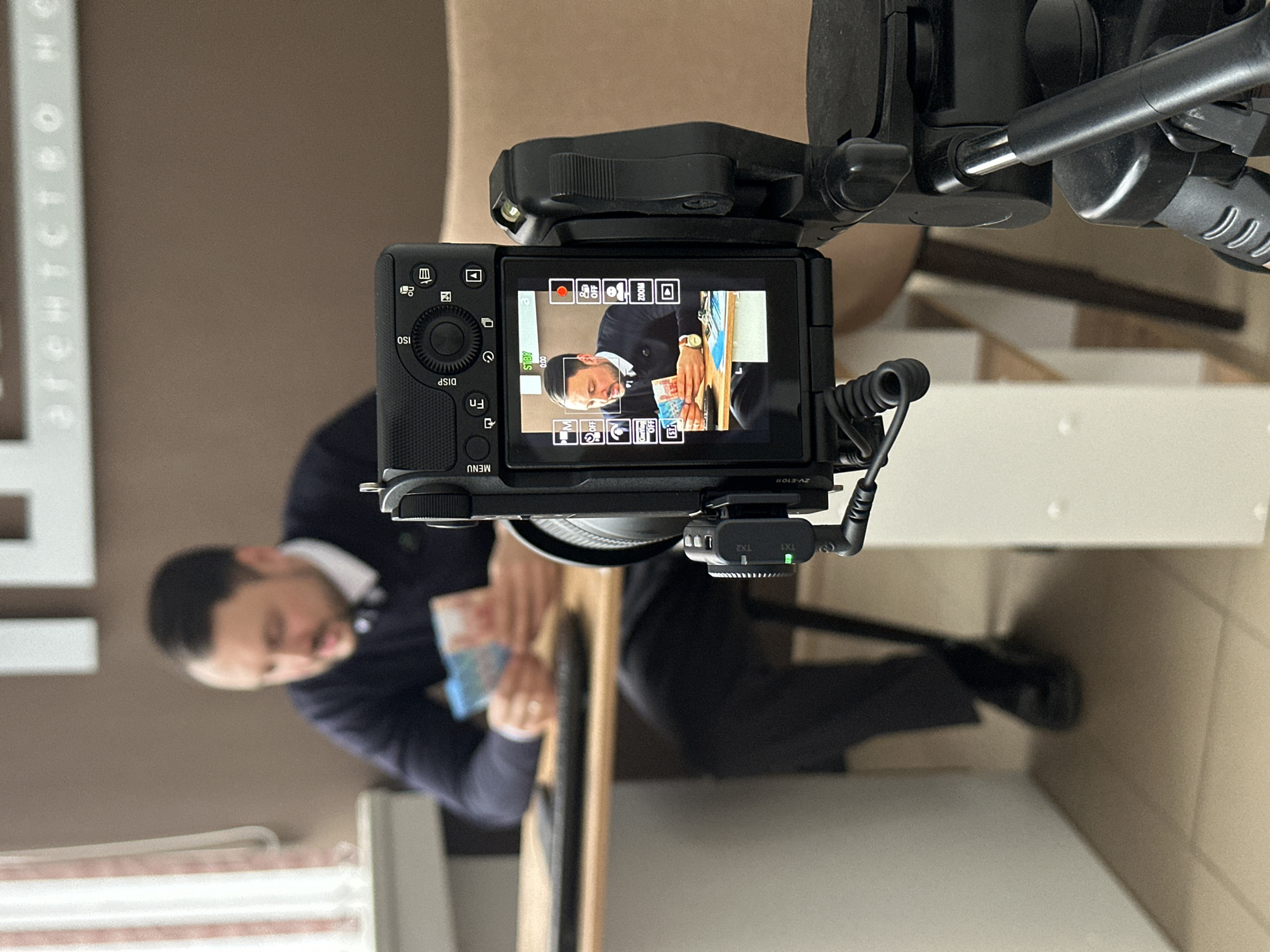 The image displays an indoor video recording setup, likely in an office or agency in Chaikovsky, Russia. The primary subject is a black Sony ZV-E10II camera, mounted vertically on a tripod or similar support system, occupying the foreground.
The camera's rear flip-out screen is open, showcasing a live view of a man. This man is also visible, albeit blurred, in the background of the main image. In the camera's display, the man is seen from the chest up, wearing a dark jacket over a white shirt. He has a beard and is looking down at his hands, where he holds a stack of banknotes. A gold watch is visible on his left wrist. The camera screen indicates "STBY" (standby mode) and "0:00", suggesting it's ready to record or paused. Several on-screen icons display camera settings, including a red recording indicator, zoom controls, and focus modes.
Attached to the camera's hot shoe is a black wireless microphone receiver, labeled "TX1" and "TX2", with a green indicator light illuminated next to "TX1". The camera body itself has visible controls and labels such as "MENU", "DISP", "Fn", and "ISO".
In the blurry background, the same man is seated behind a light-colored surface, possibly a desk, repeating the pose seen on the camera's screen. A light brown wall is behind him, with partially visible text "агентство не" (likely part of "агентство недвижимости" – real estate agency, common in Russia) printed vertically on a white panel. The overall scene suggests a professional recording or streaming session, possibly an instructional video or interview, taking place during the daytime indoors. No specific emotions are discernible from the blurred figure, but the man in the camera's view appears focused on the money.