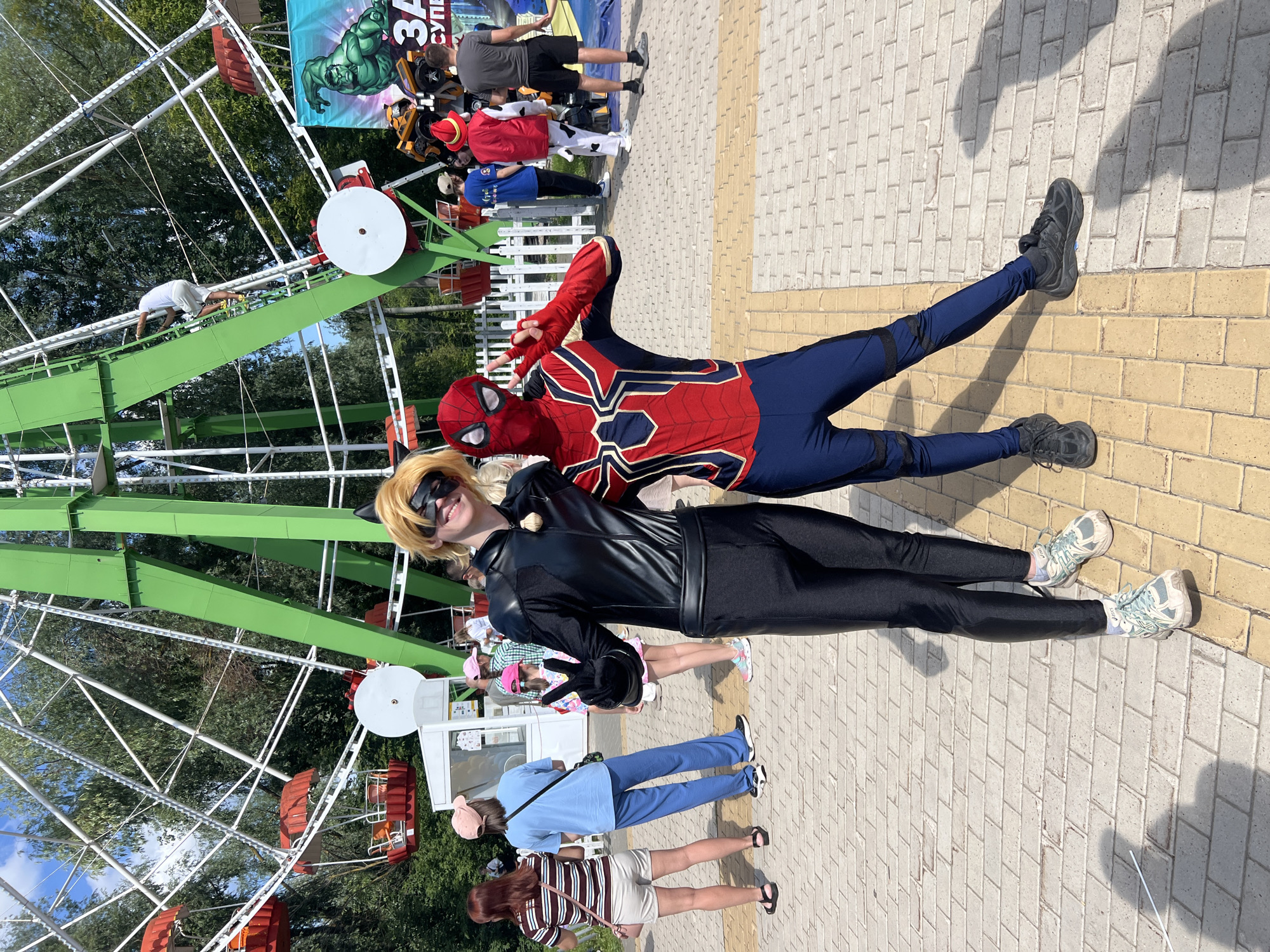 In Cheboksary, Russia, two individuals in cosplay stand on a paved walkway in front of a Ferris wheel. The person on the left is dressed as Cat Noir from Miraculous Ladybug, wearing a black catsuit, black mask with cat ears, and black gloves. They are smiling and giving a peace sign with their left hand. The person on the right is dressed as Spider-Man, in a red and blue suit with gold web detailing. They are also smiling and giving a peace sign with their right hand. In the background, the green and white structure of the Ferris wheel is prominent, with several red gondolas visible. Other people are present in the background, some appearing to be visitors at an amusement park or event. A banner featuring a depiction of the Hulk is visible to the right of the Ferris wheel. The overall environment suggests an outdoor recreational area on a sunny day.