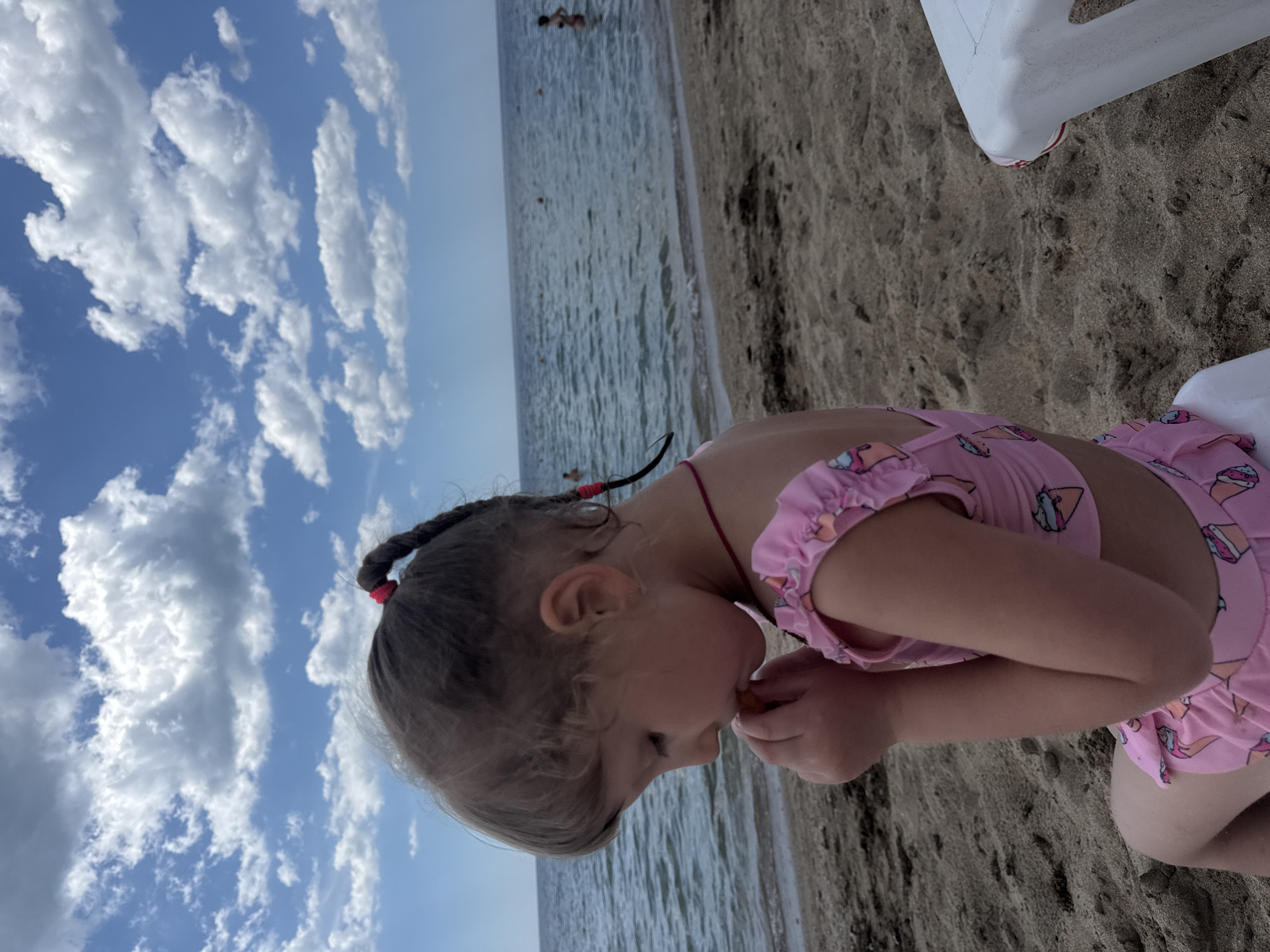 A young child sits on the sand at a beach in Yevpatoriya, Ukraine. The child is fair-skinned and wears a pink swimsuit with ice cream cone patterns. The child's hair is braided, with red bands at the ends. The child is eating something and has both hands near her mouth. The beach has light brown sand and is bordered by the sea. A white chair is partially visible in the upper right corner of the image. The sky is blue with fluffy white clouds.