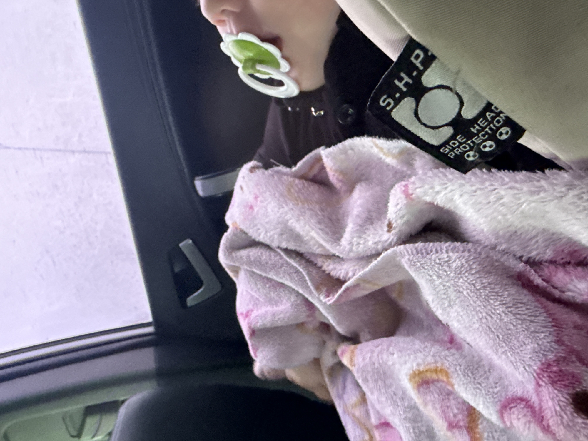 The image captures a close-up view of a baby or young child seated in what appears to be a car seat, viewed from inside a vehicle. The child's face is partially visible, showing their mouth with a green and white floral-shaped pacifier. They are wearing dark clothing, possibly a jacket or top.
A soft, pink and white patterned blanket, appearing to be plush or fleece, is draped over the child's lap and body, providing comfort. To the right of the image, a fabric tag is prominently visible, attached to what could be the car seat strap or headrest. The tag is black with white lettering and features the text "S.H.P." and "SIDE HEAD PROTECTION," along with an emblem that includes a circle and two small skull-like icons, indicating a safety feature. The car's interior is dark, with a light-colored headrest or trim visible above the tag.
Through the vehicle window on the left, an overcast or rainy sky is suggested by the diffused, light purple-grey light and subtle streaks on the glass, implying it might be daytime with poor weather. A dark car door frame or pillar is visible between the window and the child. Another dark handle or hook-like object is visible in the car interior near the window frame.
The overall scene suggests a child traveling comfortably and safely in a car, possibly in Kiselevka, Russia, given the provided location context.