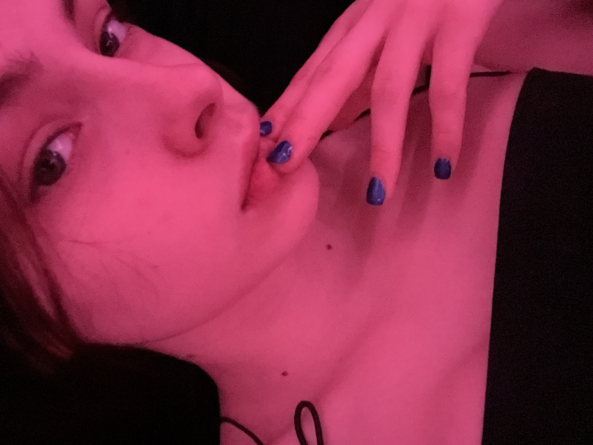 A close-up, angled portrait of a person with fair skin and short, dark hair is bathed in pink light. The subject's eyes, nose, and mouth are visible. The subject's hand is raised, with fingers near their mouth. The fingernails are painted a dark blue. The person is wearing a black top. Two moles are visible on the person's neck. The location is unknown and cannot be determined from the image.