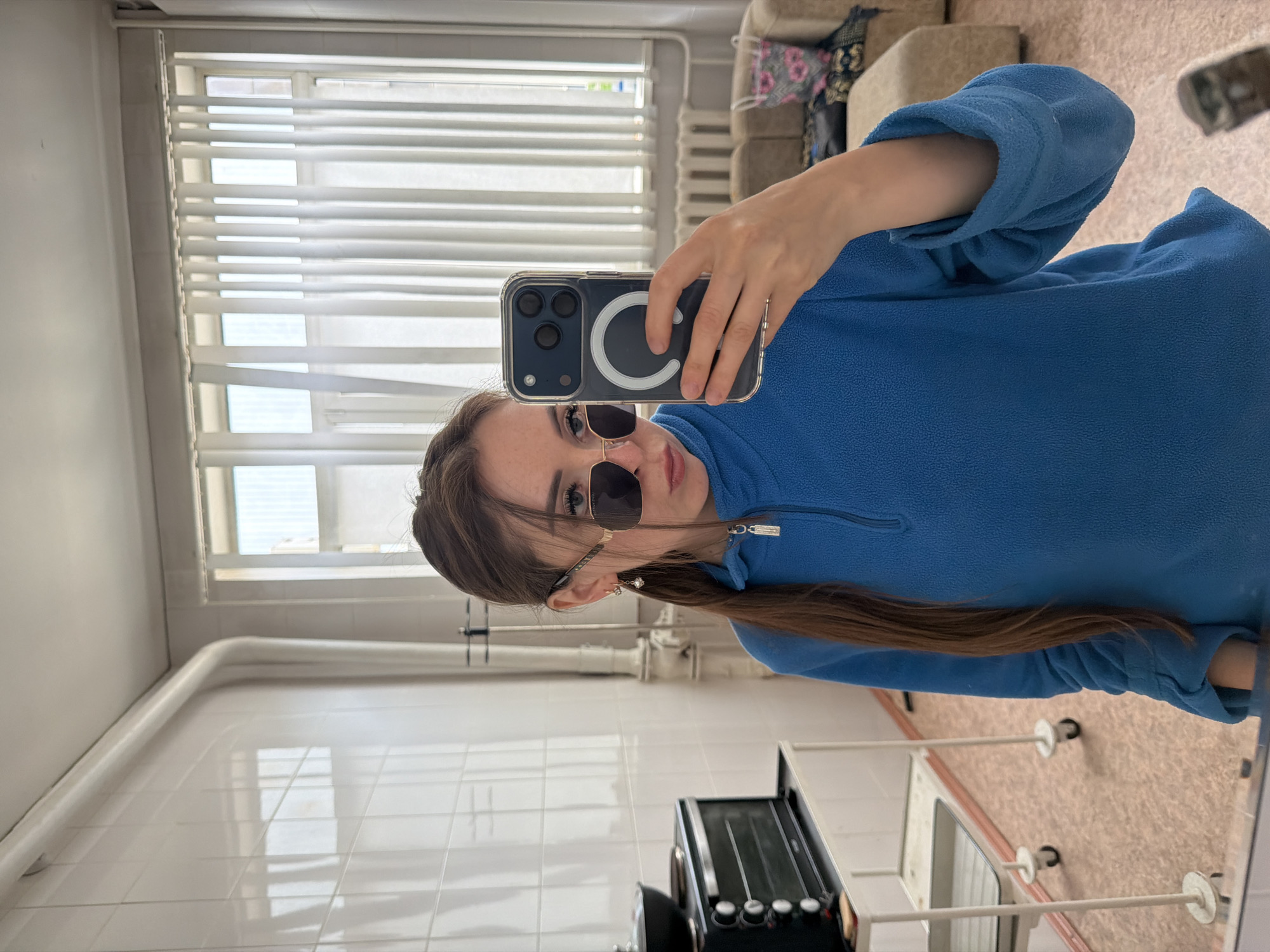A young woman with long, brown hair tied back and wearing a blue fleece jacket and sunglasses takes a selfie in a room. She is holding a smartphone in her right hand, which is positioned to capture her reflection. The smartphone has a clear case with a white ring on the back, likely for MagSafe compatibility. She has on small, pearl-like earrings. The room appears to be a domestic or institutional setting, with white tiled walls and flooring. In the background, there are windows with horizontal blinds, a radiator, and furniture such as a couch and a possible cabinet with kitchen appliances. The lighting suggests it is daytime.