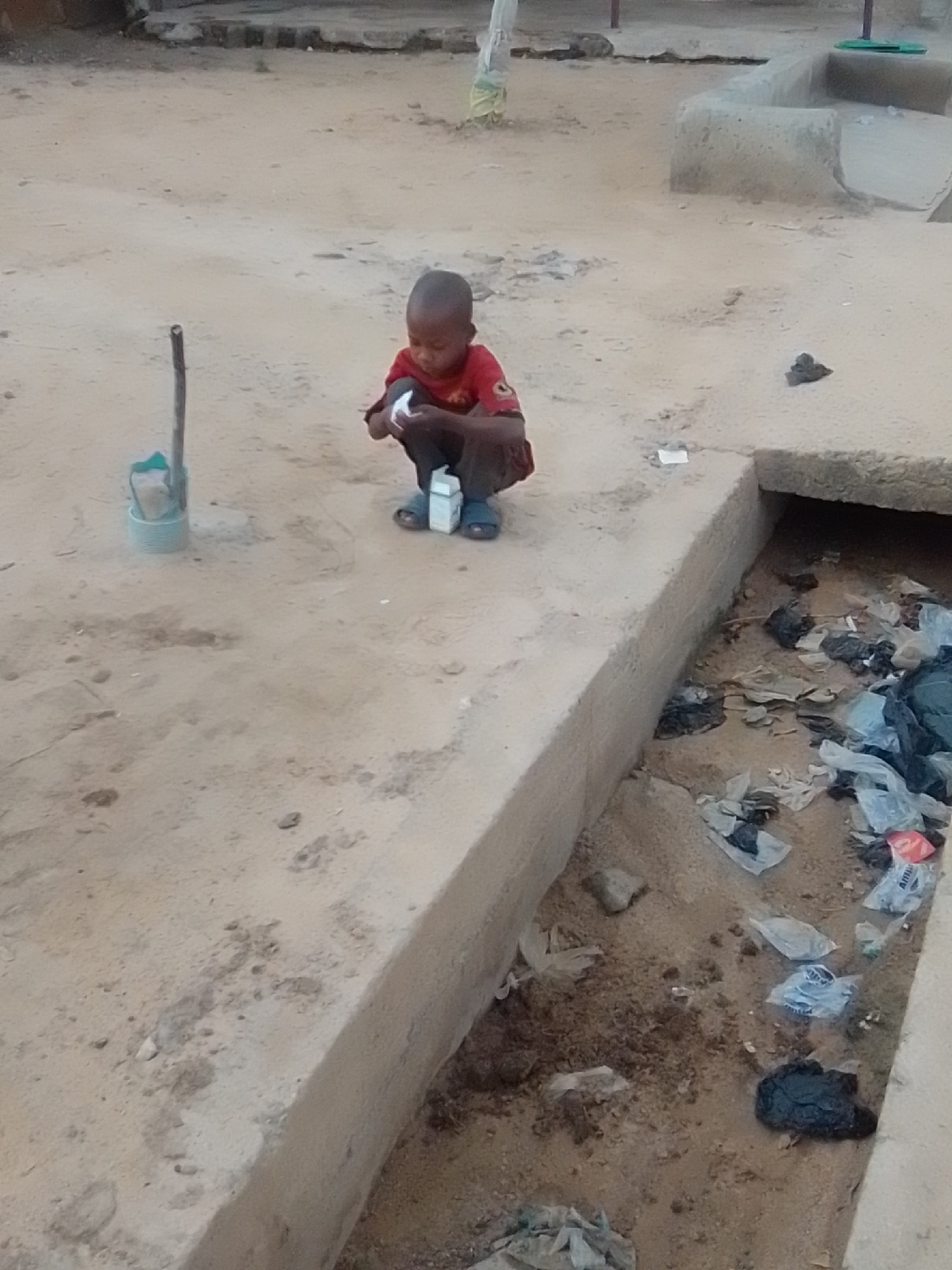 A young child in a red shirt and blue sandals is crouching on a concrete curb in Galadima, Nigeria. The child is holding a small white carton in their hands. To the left of the child, a wooden stick stands upright in a light blue bucket. The concrete curb runs alongside a drainage ditch filled with scattered plastic bags and debris. The ground surrounding the curb and ditch is dusty and uneven, with scattered pebbles and footprints. In the background, there are simple structures and sandy terrain.