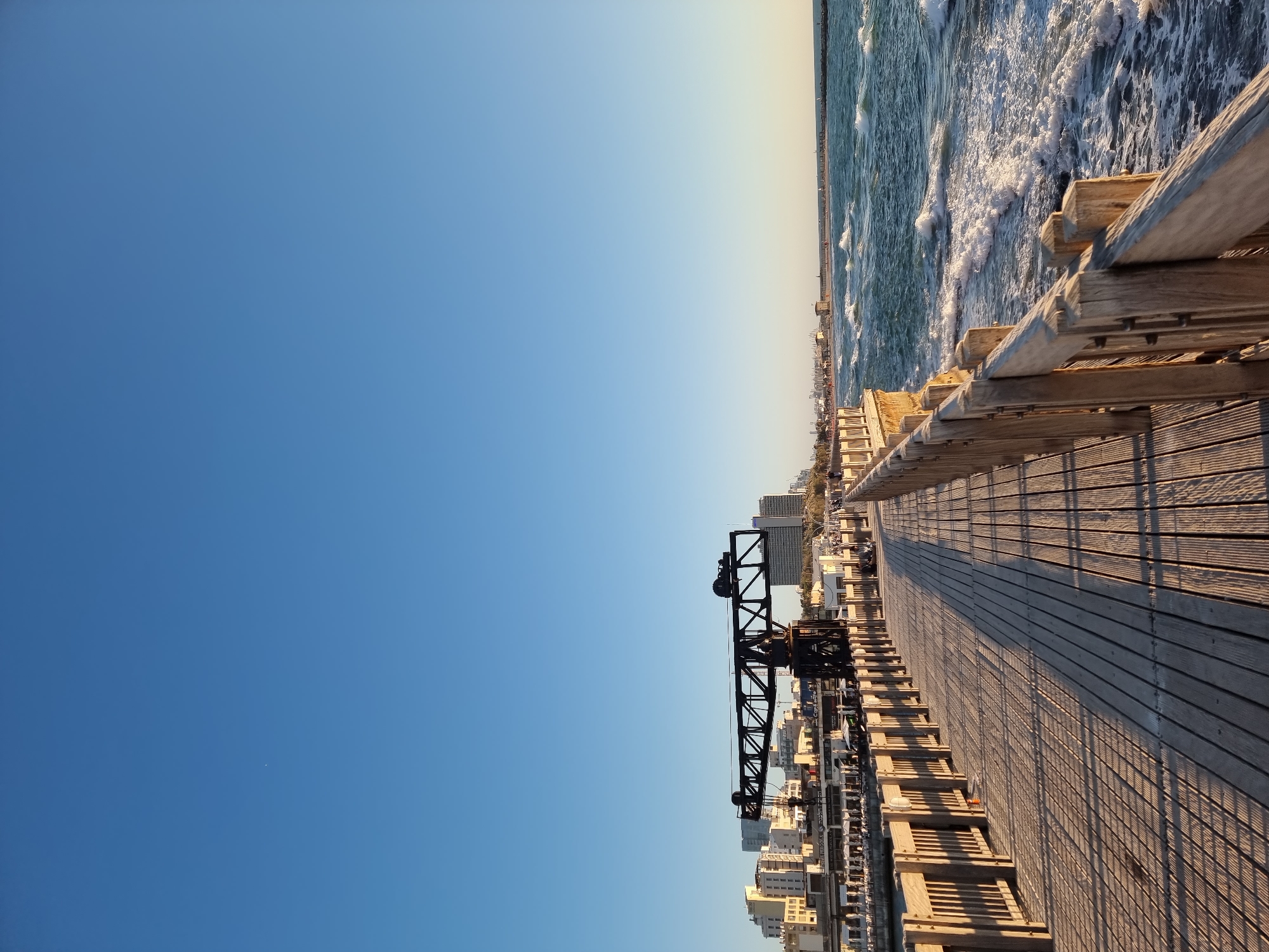 The image depicts a vibrant coastal scene, correctly oriented, showcasing a long wooden boardwalk or pier stretching into the distance under a vast, clear blue sky. The wooden planks of the boardwalk exhibit detailed texture and are marked by strong, elongated shadows, indicating a low sun angle typical of late afternoon or early morning.
To the right of the boardwalk, the sea unfolds with energetic white-capped waves crashing, suggesting a lively ocean. A robust wooden railing runs alongside the boardwalk, separating it from the water. On the left side, a cityscape is visible, featuring various buildings, many of which appear modern and light-colored. A striking black, metallic gantry-like or crane structure, with a distinctive truss design, stands prominently above the buildings, hinting at the area's industrial past.
No people or distinct activities are clearly visible, conveying a sense of openness and calm. The weather is bright and sunny, with no clouds in the sky. This scene is characteristic of the revitalized Tel Aviv Port in Tel Aviv, Israel, where historical port elements blend with contemporary leisure spaces. There is no visible text legible in the image.