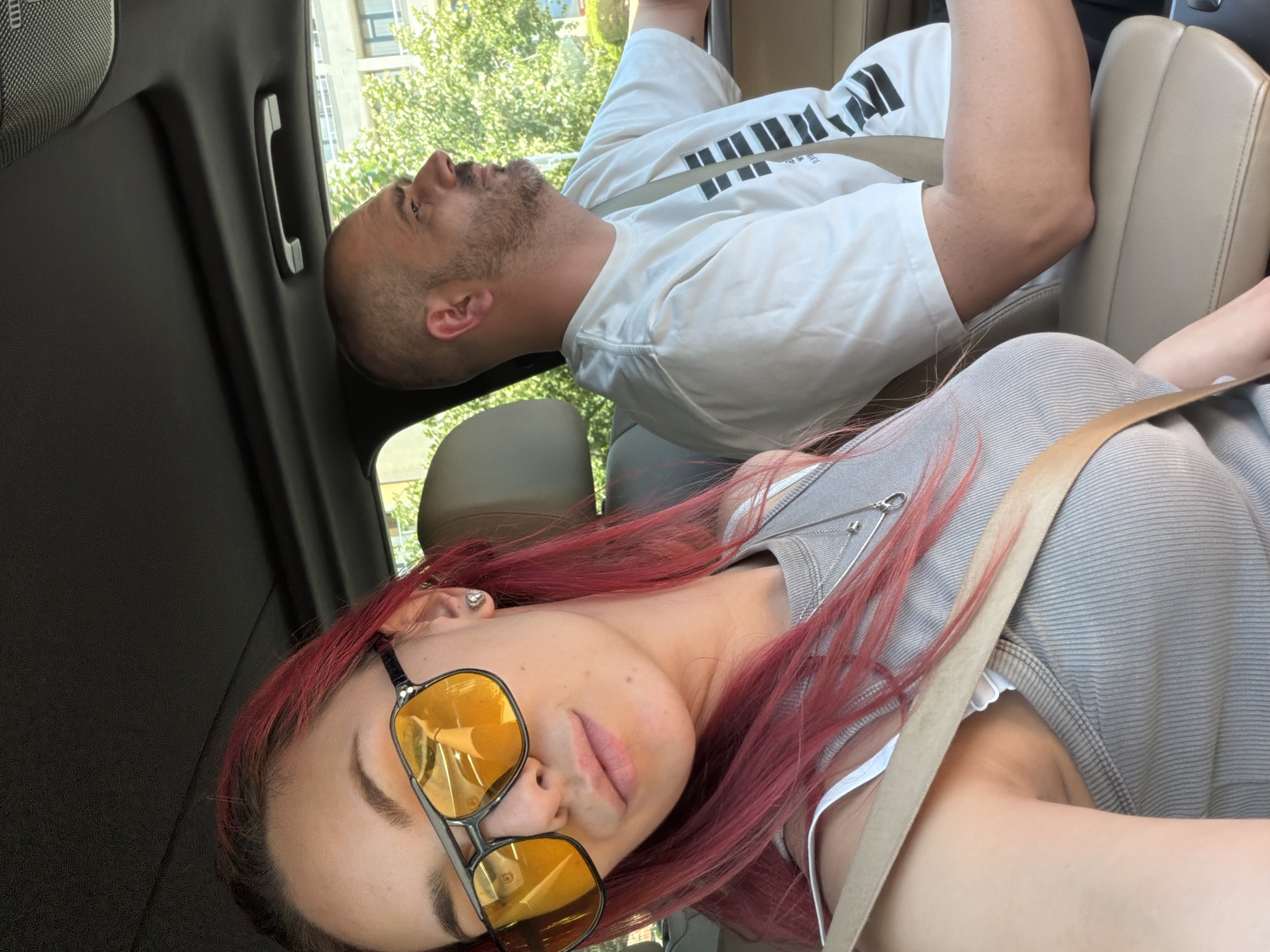 In the city of Nesebar, Bulgaria, a man and a woman are inside a car. The woman, with bright pink hair and yellow-tinted sunglasses, is positioned in the foreground, taking a selfie. She is wearing a grey ribbed tank top and a white bra. A beige seatbelt is across her chest. The man, with a shaved head and a beard, is in the driver's seat to her right, looking out the window. He is wearing a white t-shirt with a black graphic design. The car's interior, with beige leather seats, is visible, as is the lush greenery and buildings outside the windows, suggesting a sunny day in an urban or suburban environment.