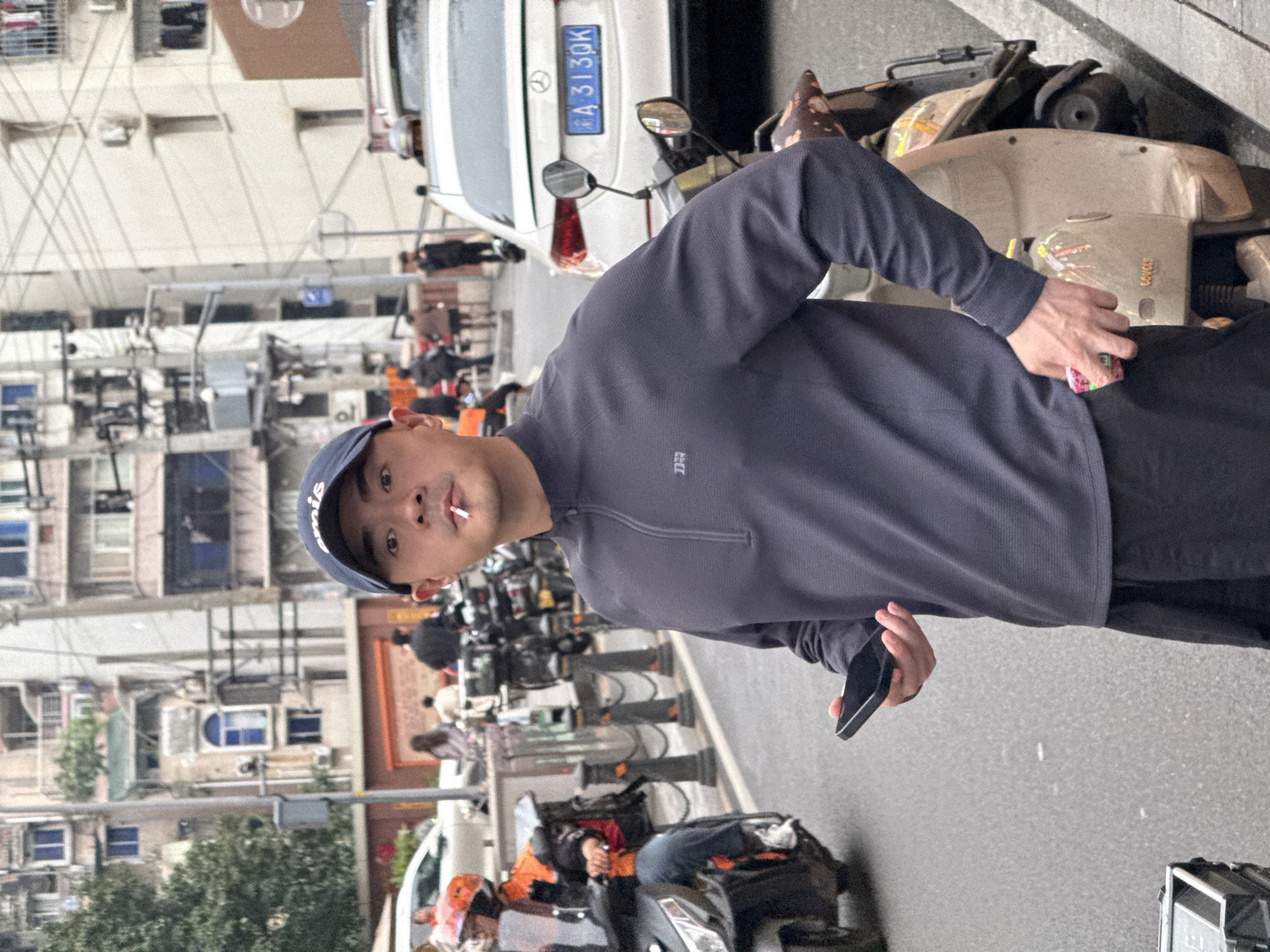A street scene in Liangjiang, China. A man in a dark blue long-sleeved shirt and a blue baseball cap is holding a smartphone in his right hand and appears to have a cigarette in his mouth. He is standing near a white car with the license plate A43130K. Several motorcycles are parked along the street. Buildings line the left side of the frame.