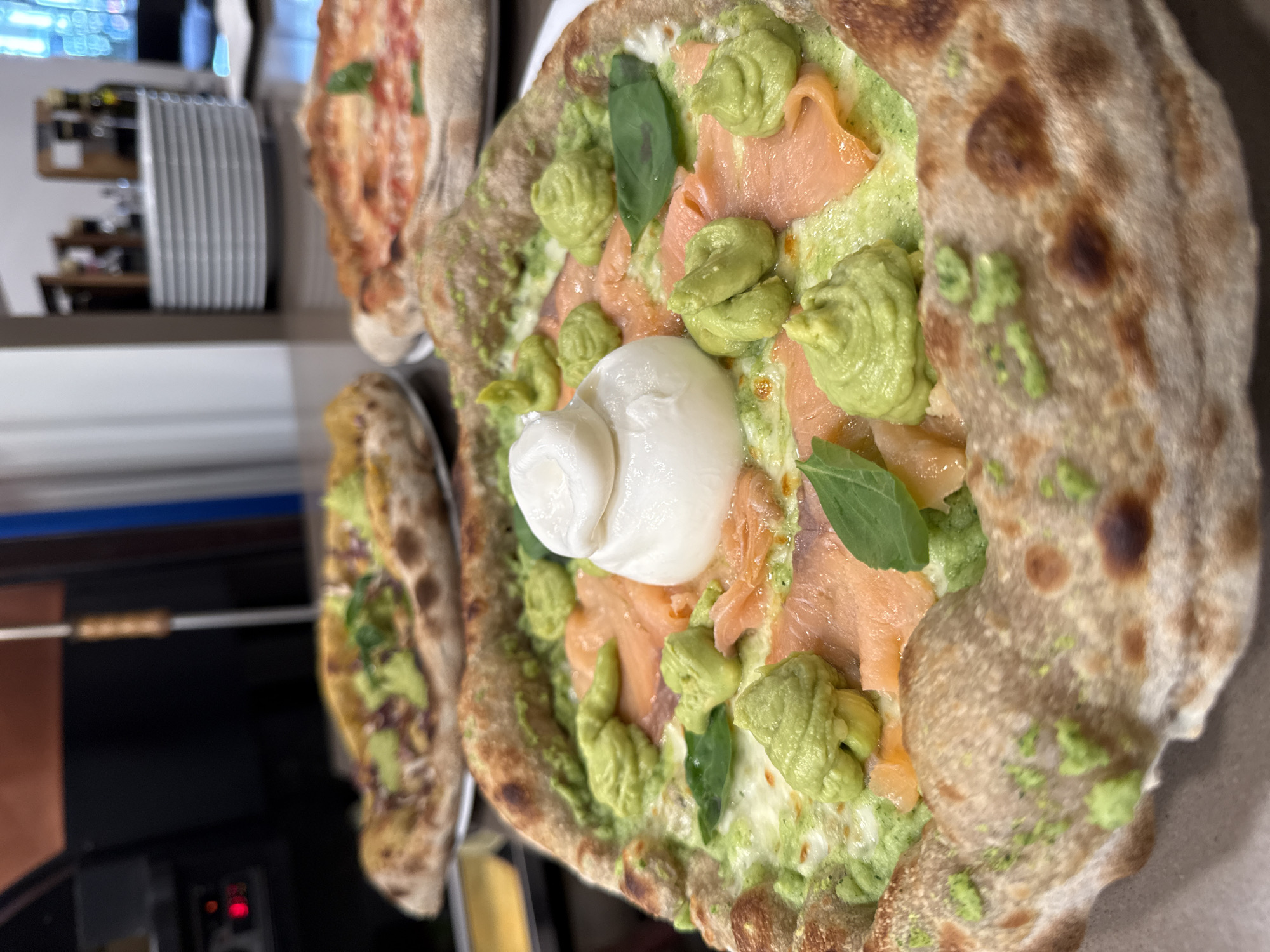 A close-up shot features a close-up of a gourmet pizza with smoked salmon, burrata cheese, and a green sauce, likely avocado-based, with dollops of the sauce strategically placed around the pizza. The pizza has a thick, artisanal crust with some charring, indicating it was likely cooked in a wood-fired oven. Two other pizzas are visible in the background, slightly out of focus, one topped with a red sauce and cheese, and another with green toppings. The pizzas are presented on plates on a countertop. In the background, a stack of white plates and a glimpse of kitchen equipment can be seen, suggesting the setting is a restaurant kitchen or a pizzeria. The image is well-lit, highlighting the textures and colors of the food. The specific location in Trento, Italy, is not identifiable from the image itself.