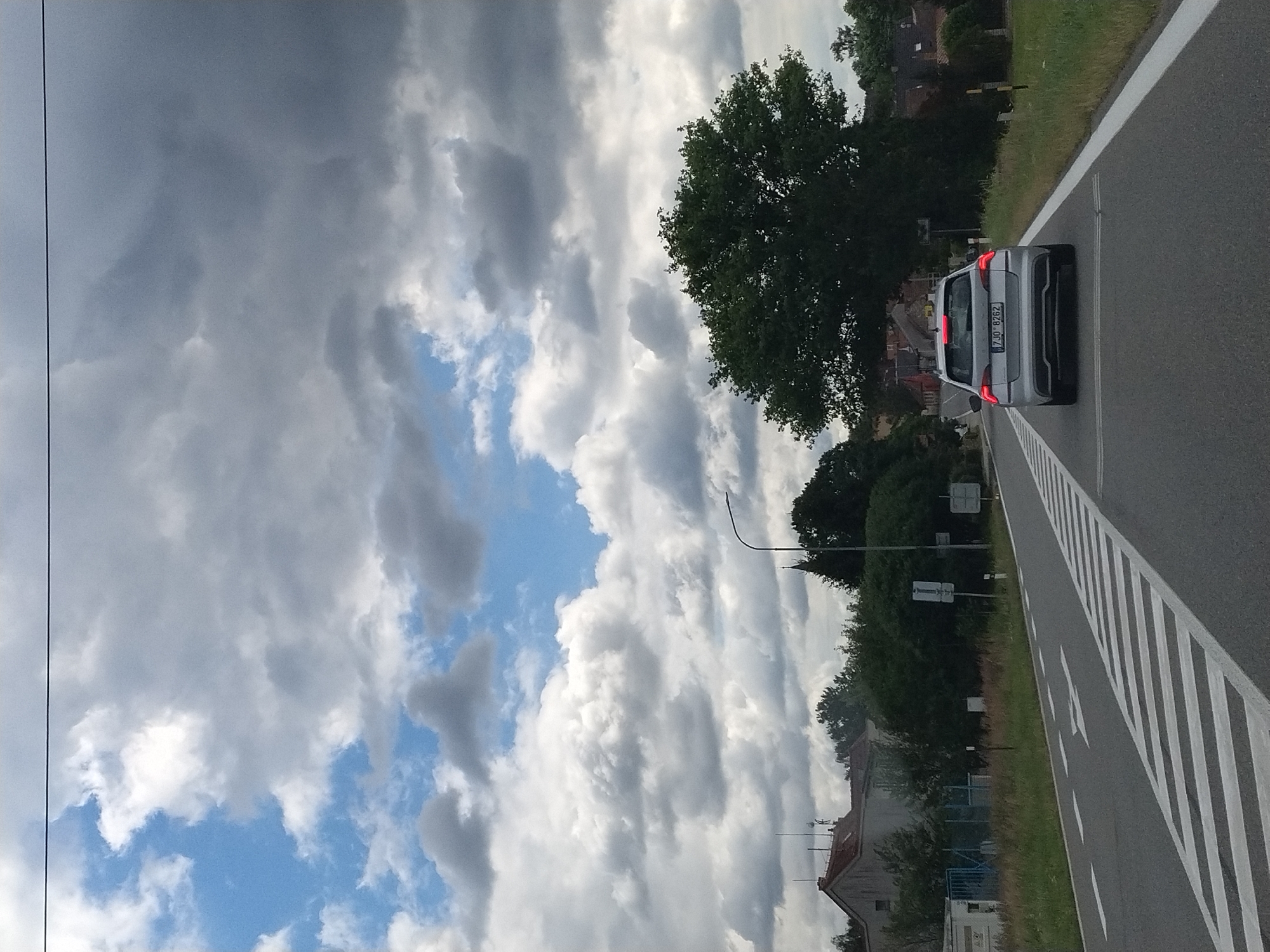 A silver Skoda Octavia is driving on a paved road in Bernartice, Czechia, on a cloudy day. The car has a Czech license plate that reads "7JO 8262". The road has white lane markings and a pedestrian crossing in the foreground. On the left side of the road, there are some buildings and trees. On the right side of the road, there is a grassy verge and trees. In the background, there are more buildings and trees. The sky is filled with dramatic clouds.