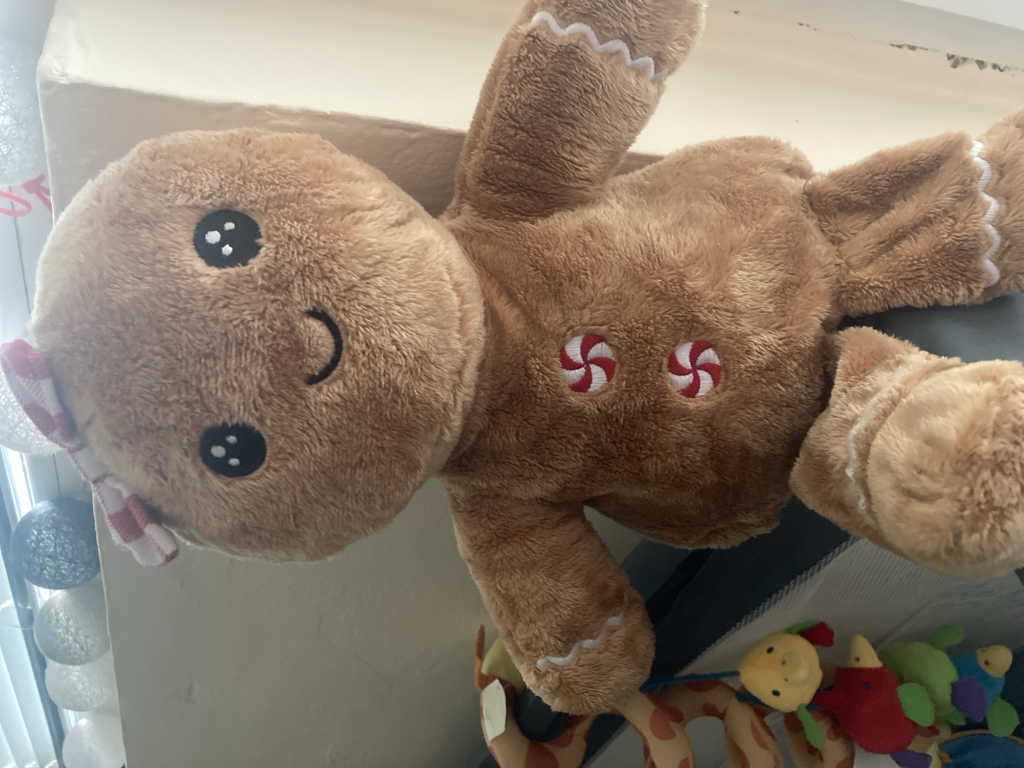 A plush gingerbread man toy is centered in the frame, facing forward. It has a brown, furry texture with embroidered black eyes and a smiling mouth. Two red and white swirled candy designs are sewn onto its chest, and white ric-rac trim adorns its arms and legs. A red and white checkered ribbon is tied on its head. To the left of the gingerbread man, a baby toy with multiple colorful animal characters hanging from a string is visible. In the background, decorative white and gray spherical lights are strung across a windowsill. The scene appears to be indoors, likely a domestic setting in Burgas, Bulgaria, though this specific location cannot be definitively determined from the image alone.