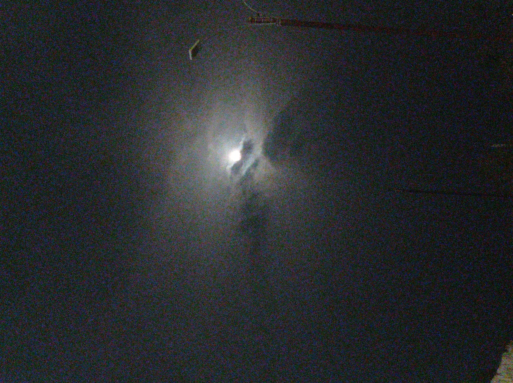 The image captures a dark, heavily pixelated night scene, characterized by significant visual noise, indicating very low light conditions. At the center, a bright, diffused light source, strongly resembling the moon, is visible through what appears to be thick mist, clouds, or fog. This light creates a prominent, soft glow and halo effect, with swirling patterns of obscuring atmospheric elements around it.
No people are visible. In the upper left portion of the frame, a small, light-colored, elongated object is partially discernible, possibly suspended. Towards the upper right, a dark, horizontal, elongated structure, consistent with a beam, pipe, or electrical wiring, stretches across the top of the image. These objects suggest an outdoor setting, potentially urban or semi-urban, where overhead structures are present against the night sky.
The overall scene depicts a hazy or foggy night in Porlamar, Venezuela, with the moon partially obscured. No visible text is present.