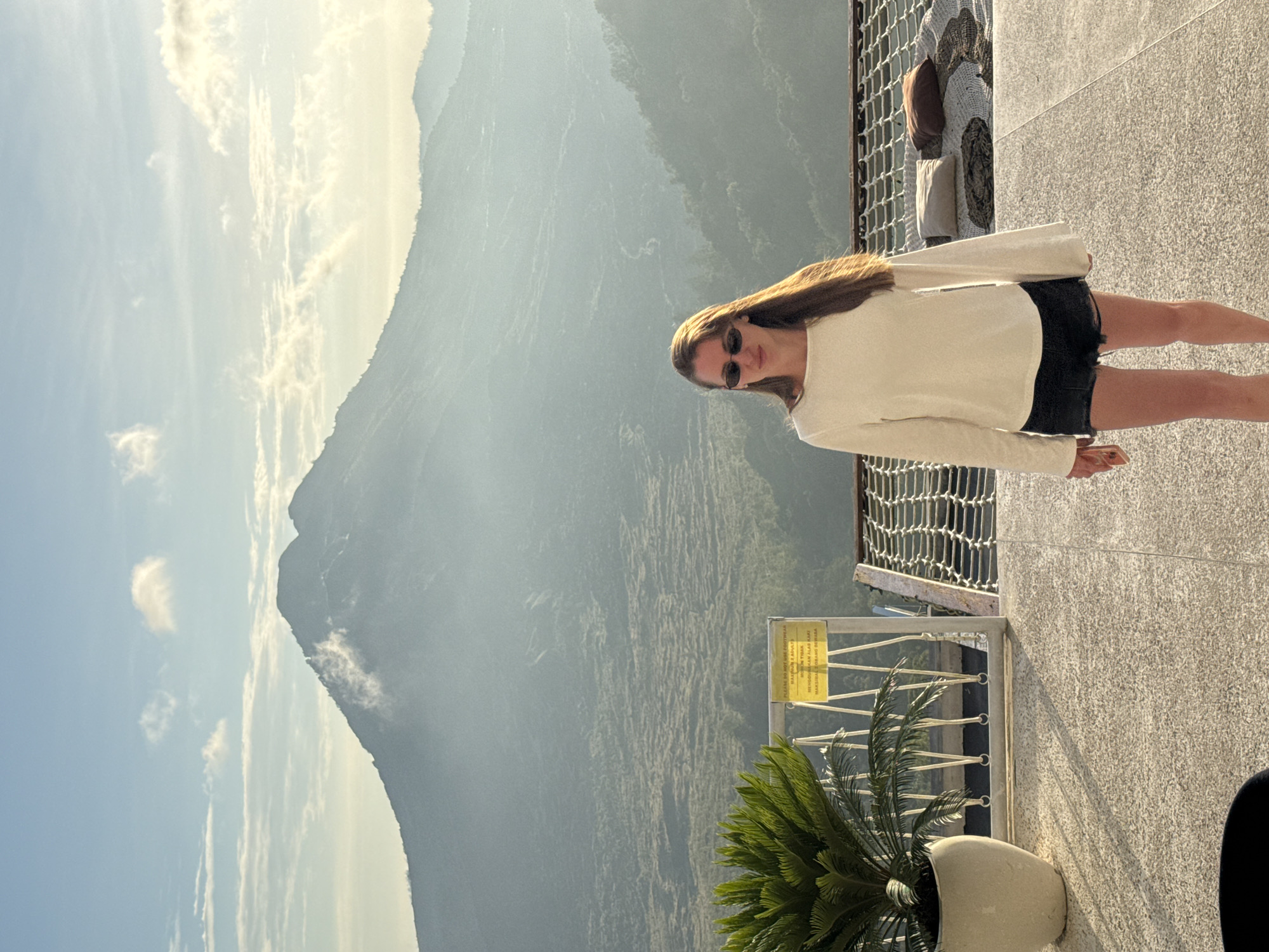 A long-haired woman wearing sunglasses, a light-colored long-sleeved shirt, and dark shorts stands on a concrete surface with a metal safety railing. Behind her is a mountainous landscape, possibly in the region of Kedisan, Indonesia. In the foreground, a potted plant sits near a sign with writing on it. A net-like structure is visible behind the woman. The sky is partly cloudy.