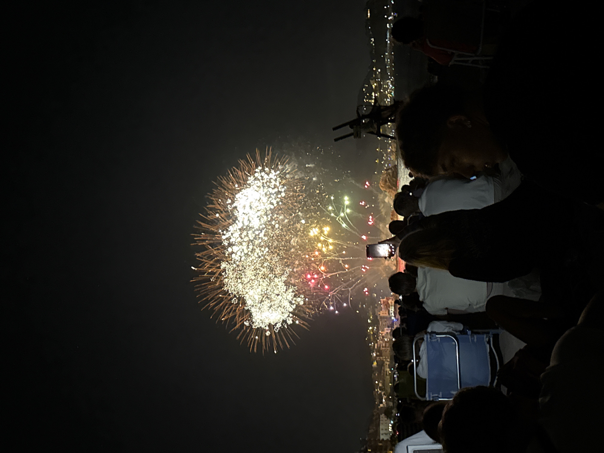 In Blanes, Spain, during a nighttime event, a crowd of spectators is gathered on a beach to watch a vibrant fireworks display over the water. The sky is illuminated by a large burst of white and gold fireworks, with colorful sparks of red, yellow, and green radiating outwards. In the foreground, silhouetted figures of people are seated on the sand, some in chairs, facing the spectacle. A few individuals are holding up smartphones to capture the moment. The background reveals the distant lights of the city of Blanes, nestled along the coastline, with buildings and hills visible. The overall scene depicts a festive atmosphere during a fireworks show in a coastal Spanish city.