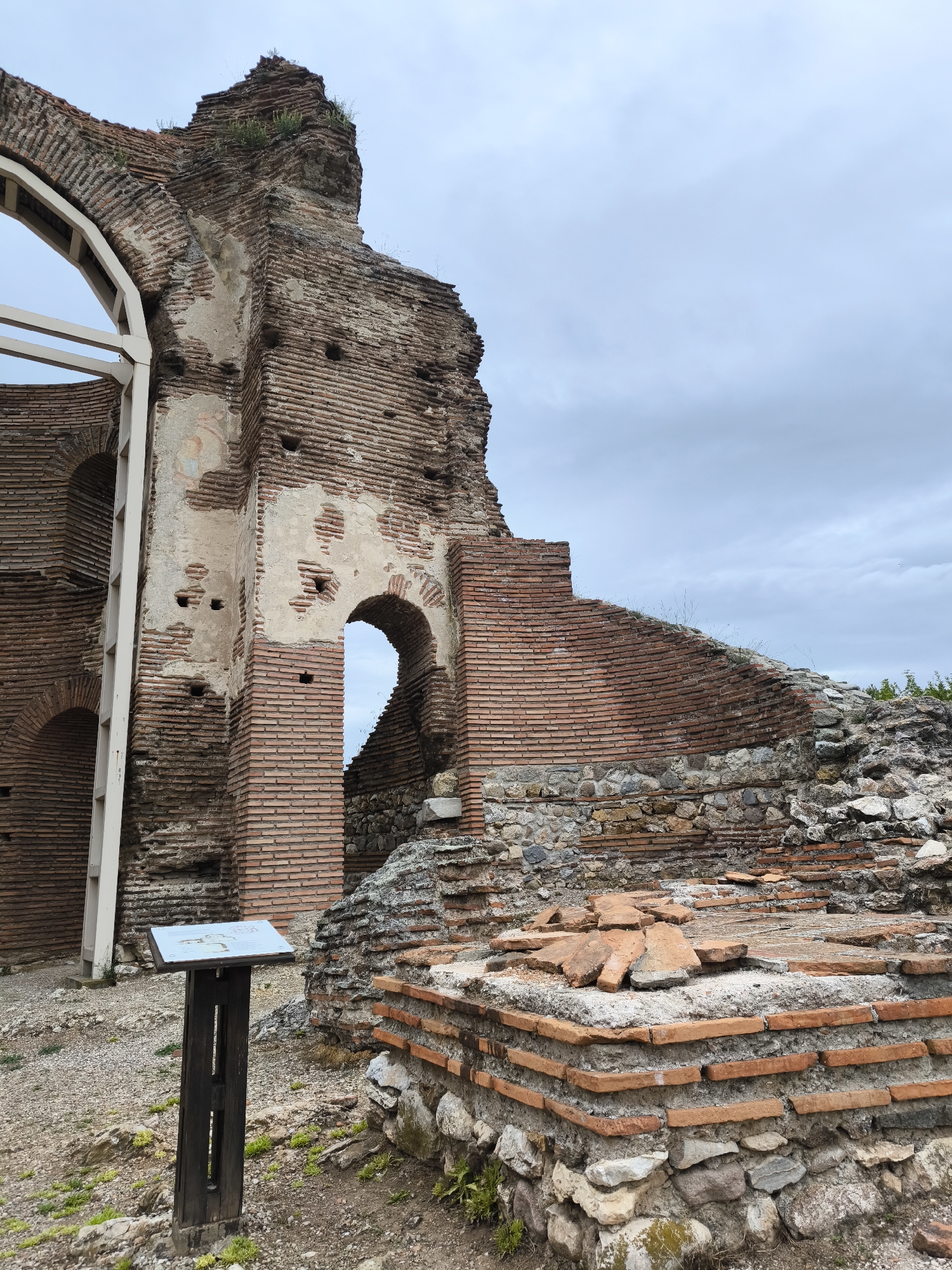 The image depicts a section of the ruins of the Red Church, an early Christian basilica located near Perushtitsa, Bulgaria. The ruins consist of red brick walls with arched openings, some filled in with stone. A partial arch with modern scaffolding provides structural support. A wooden information sign is visible in the foreground. Debris of broken bricks and stones are scattered on the ground. The sky is overcast. No people are visible.