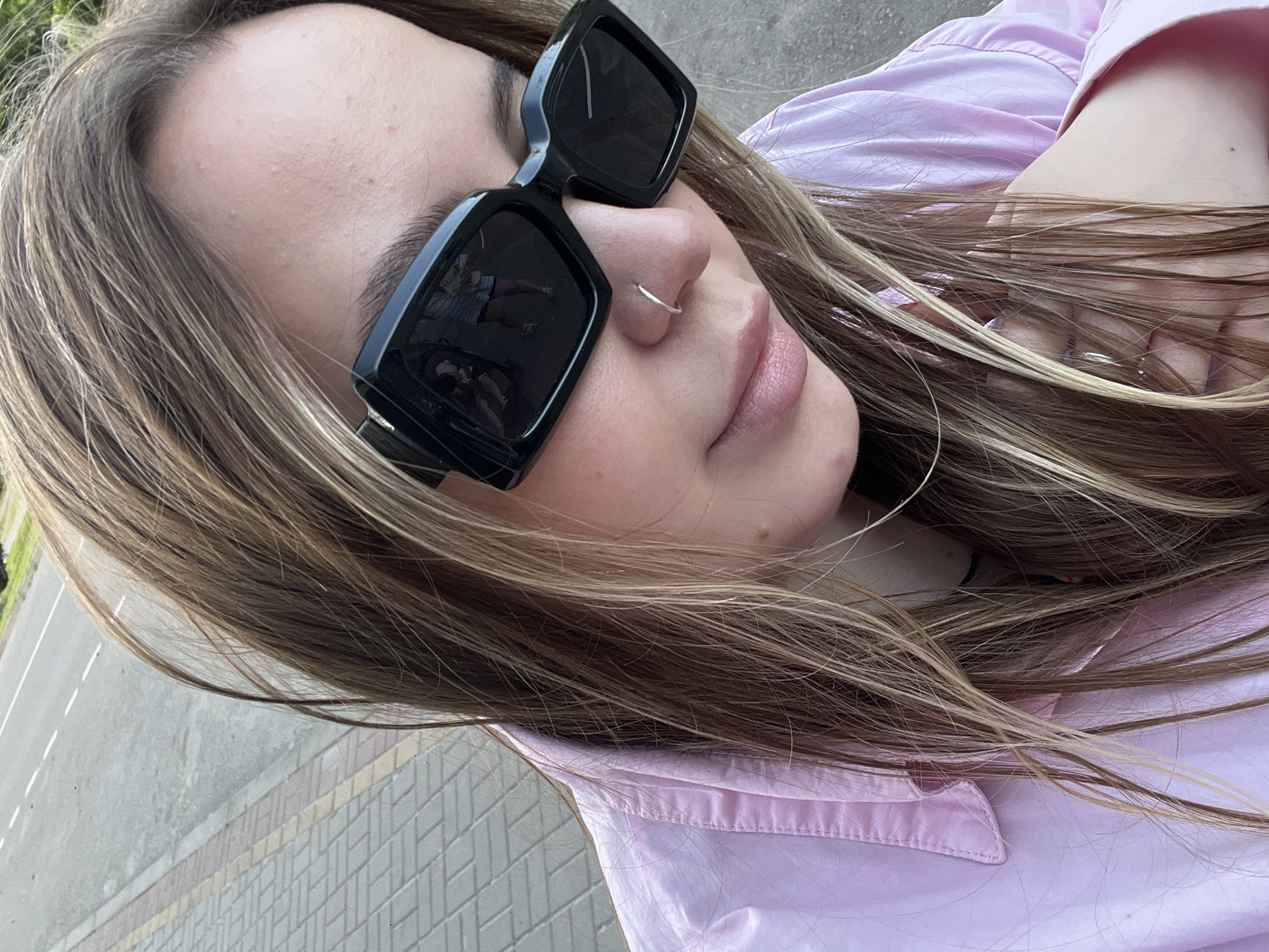 A young woman with long, light brown hair with blonde highlights is wearing black, square-framed sunglasses and a silver nose ring. She is wearing a light pink collared shirt. The photo appears to have been taken outdoors, with a sidewalk visible in the background. Based on the prompt, this is located in the city of Novopokrovskaya, Russia.